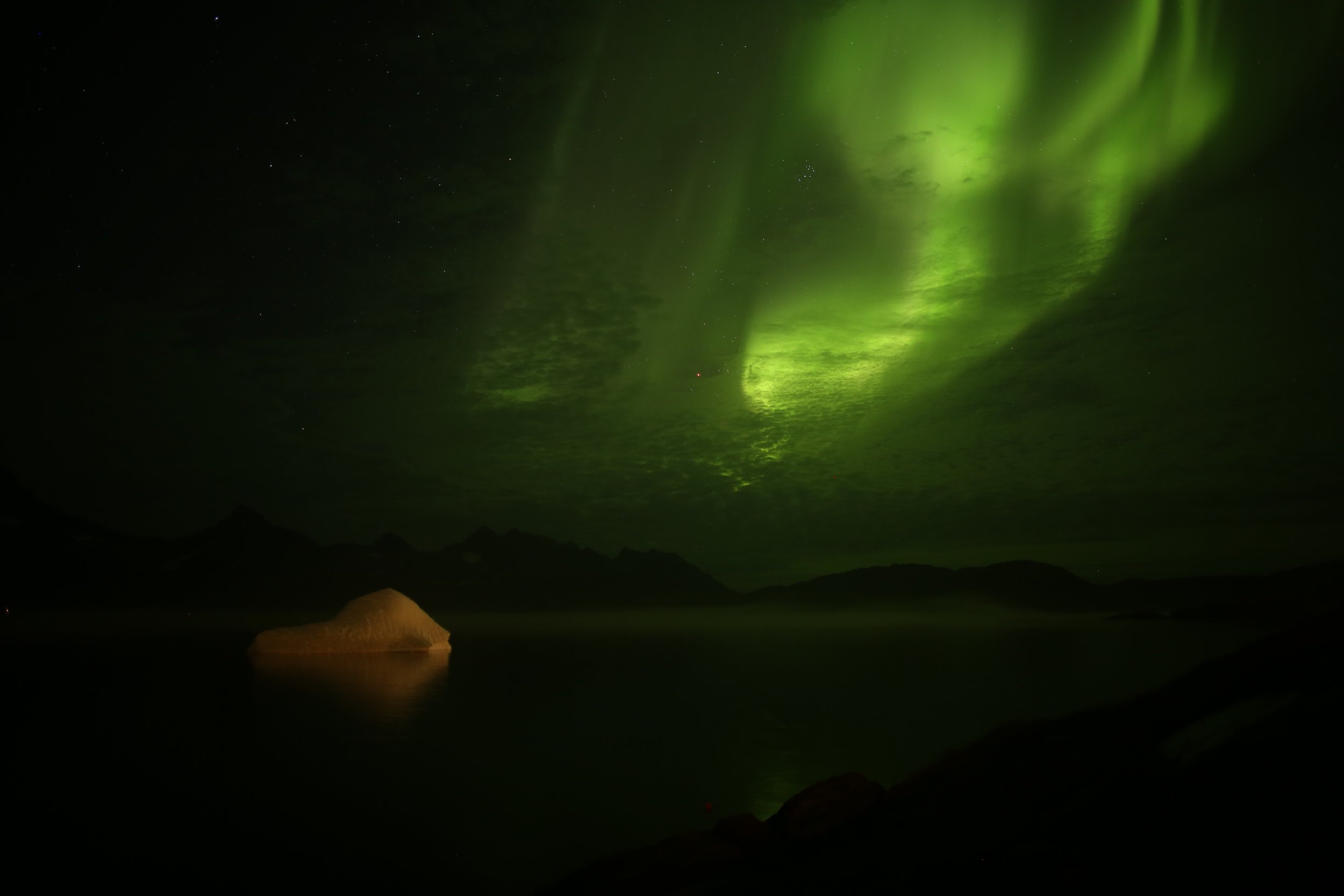 Northern lights illuminating the night sky over a mountain lake with a large rock in the foreground.