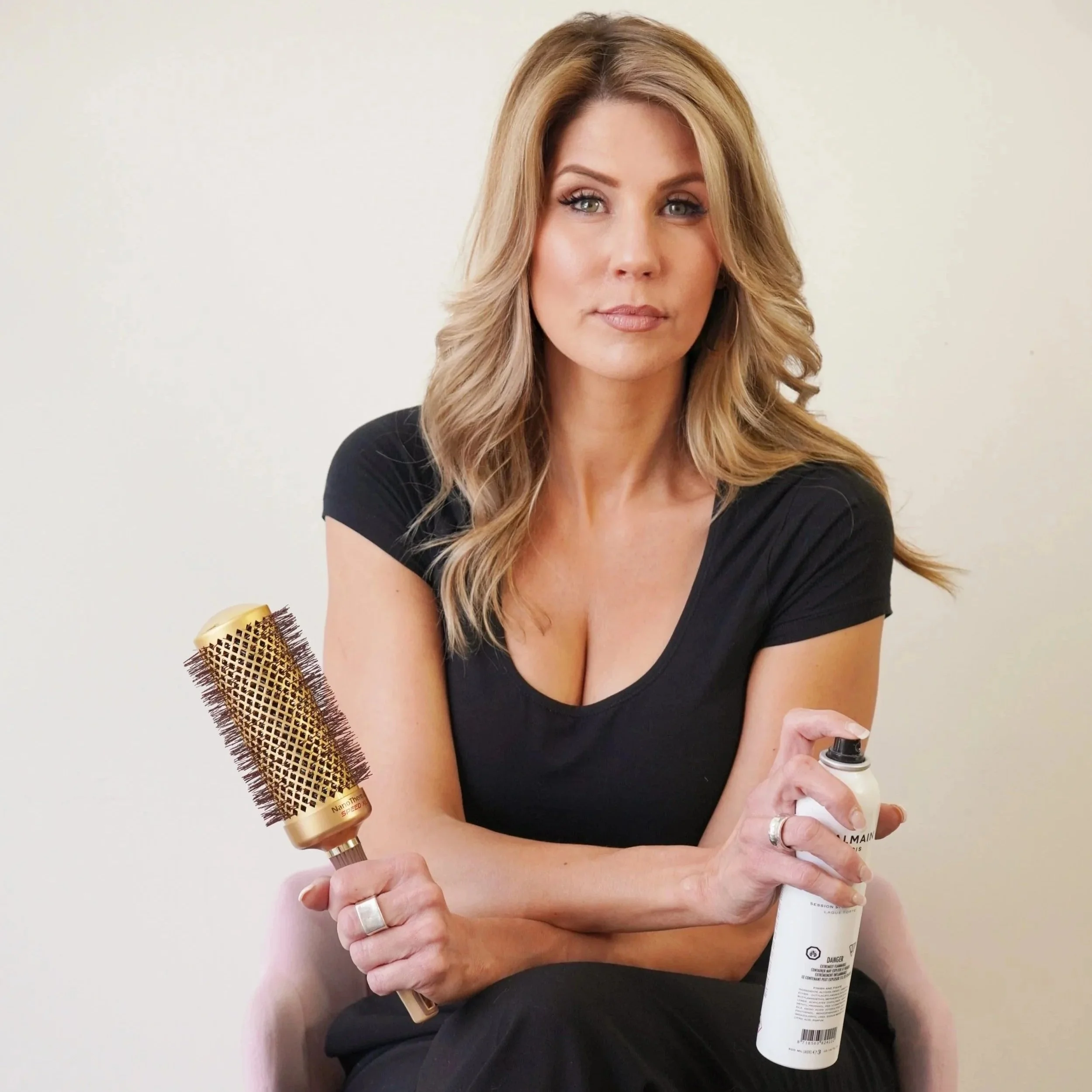 A woman with blonde hair wearing a black top, holding a bottle of lotion or cream in one hand and a hairbrush in the other, against a plain light-colored background.