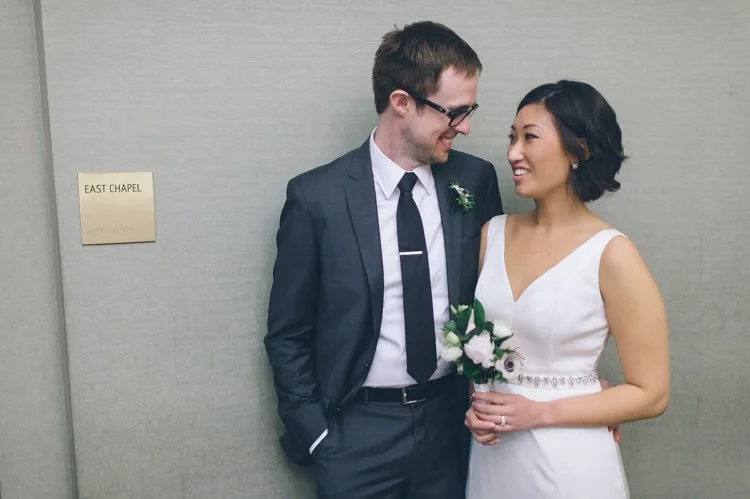 Bride and groom smiling at each other near a sign that reads 'East Chapel'. The groom is in a suit, and the bride is holding a bouquet, wearing a white dress.