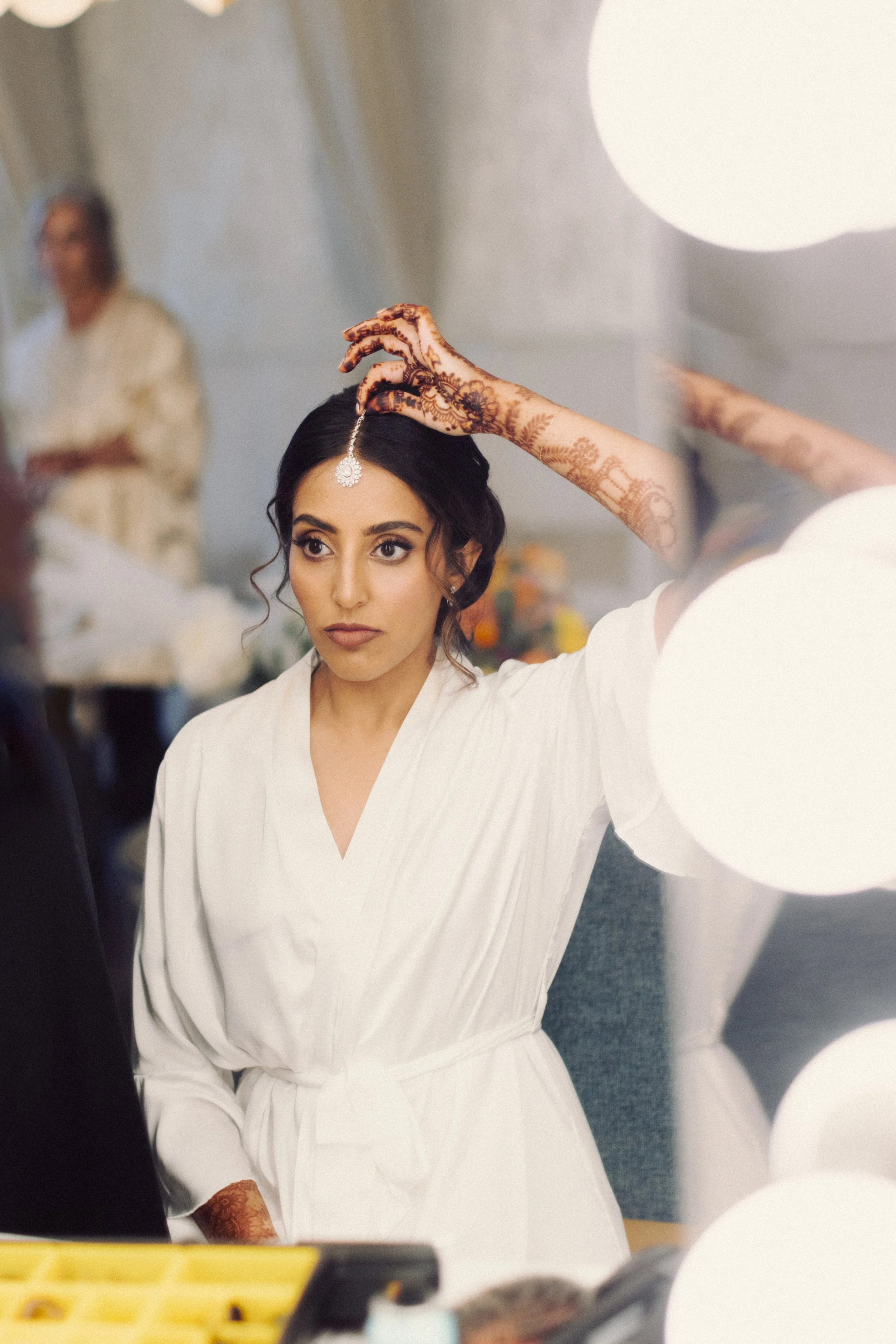 Woman in white robe applying jewelry in front of a mirror, with henna designs on hands.
