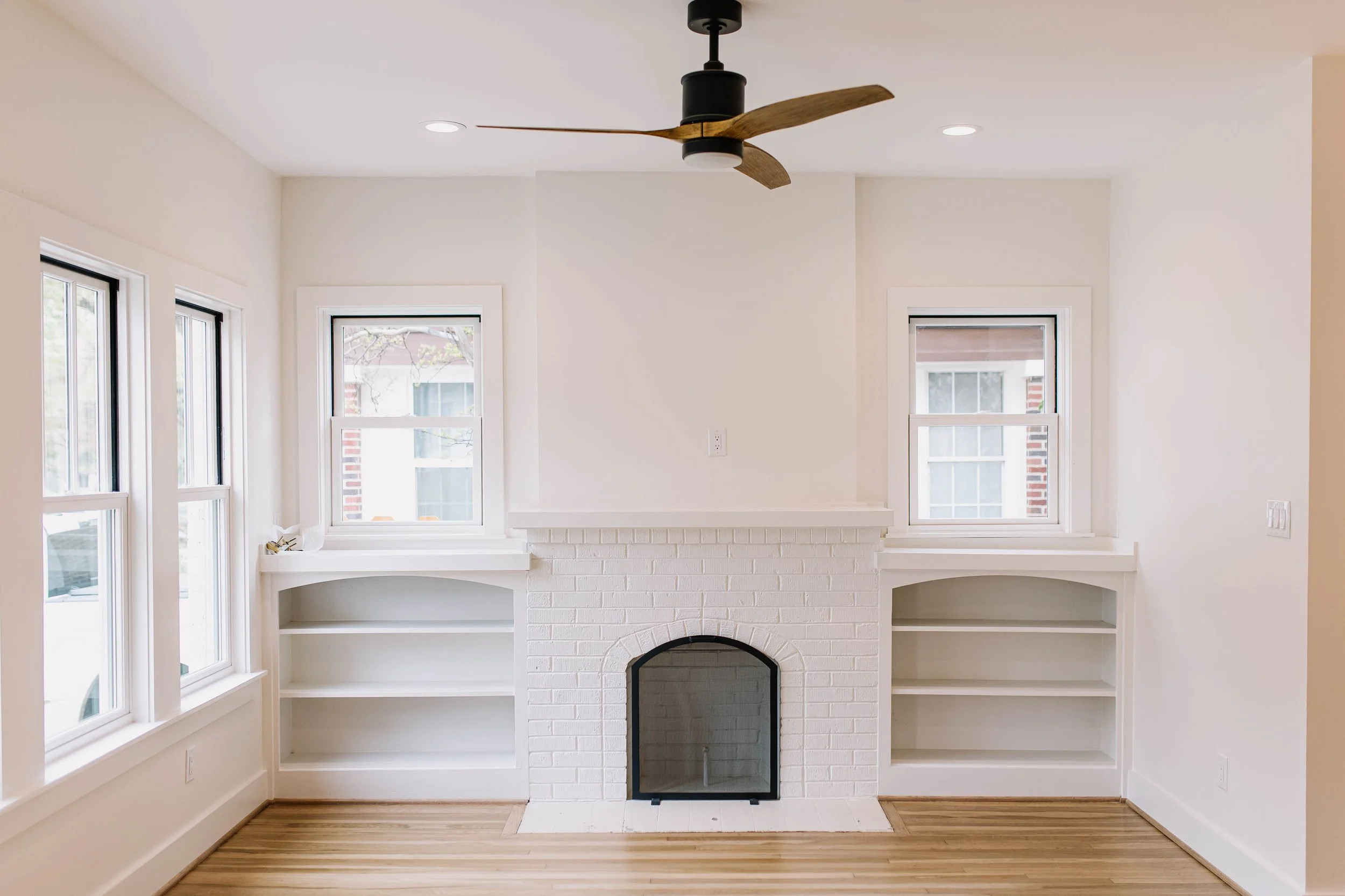 Living room with white brick fireplace, built-in shelves, three windows, ceiling fan, and hardwood floors.