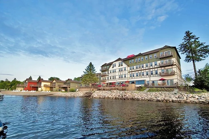 Lakeside hotel or apartment complex with multiple stories, balconies, and outdoor seating, adjacent to a body of water with a rocky shoreline and neighboring small buildings, under a partly cloudy sky.