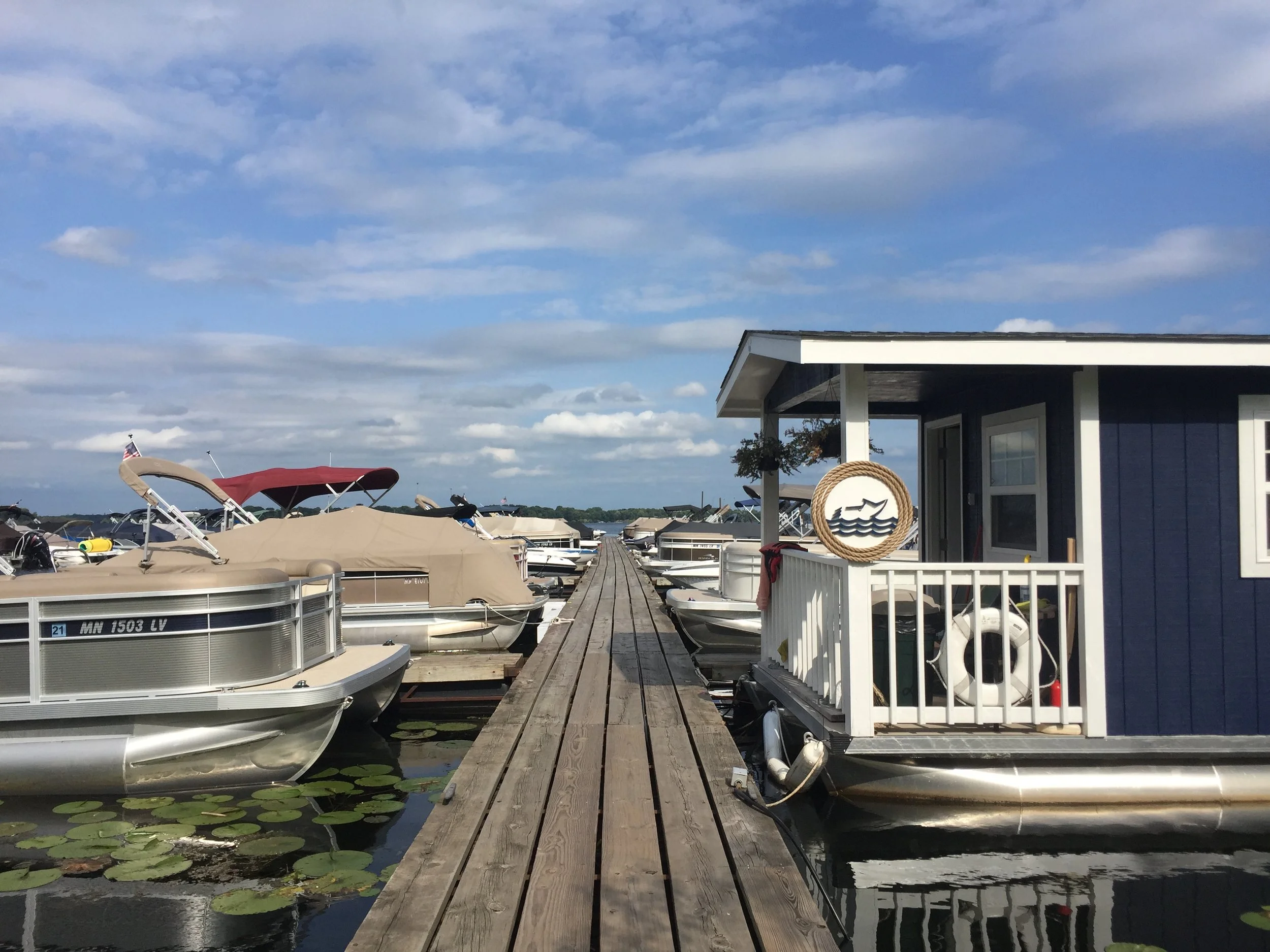 Dock leading to boats and houseboat at a marina under a blue sky with clouds.