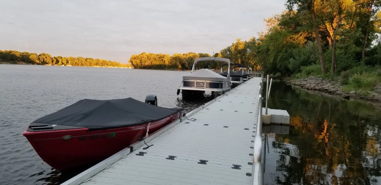 Dock with boats on a calm river, surrounded by trees with fall foliage, during sunset.
