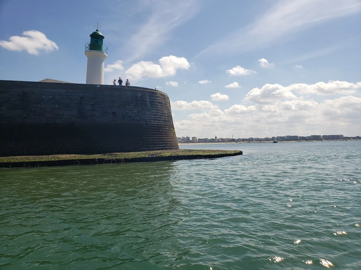 View of a lighthouse on a pier by the sea with a city skyline in the background.
