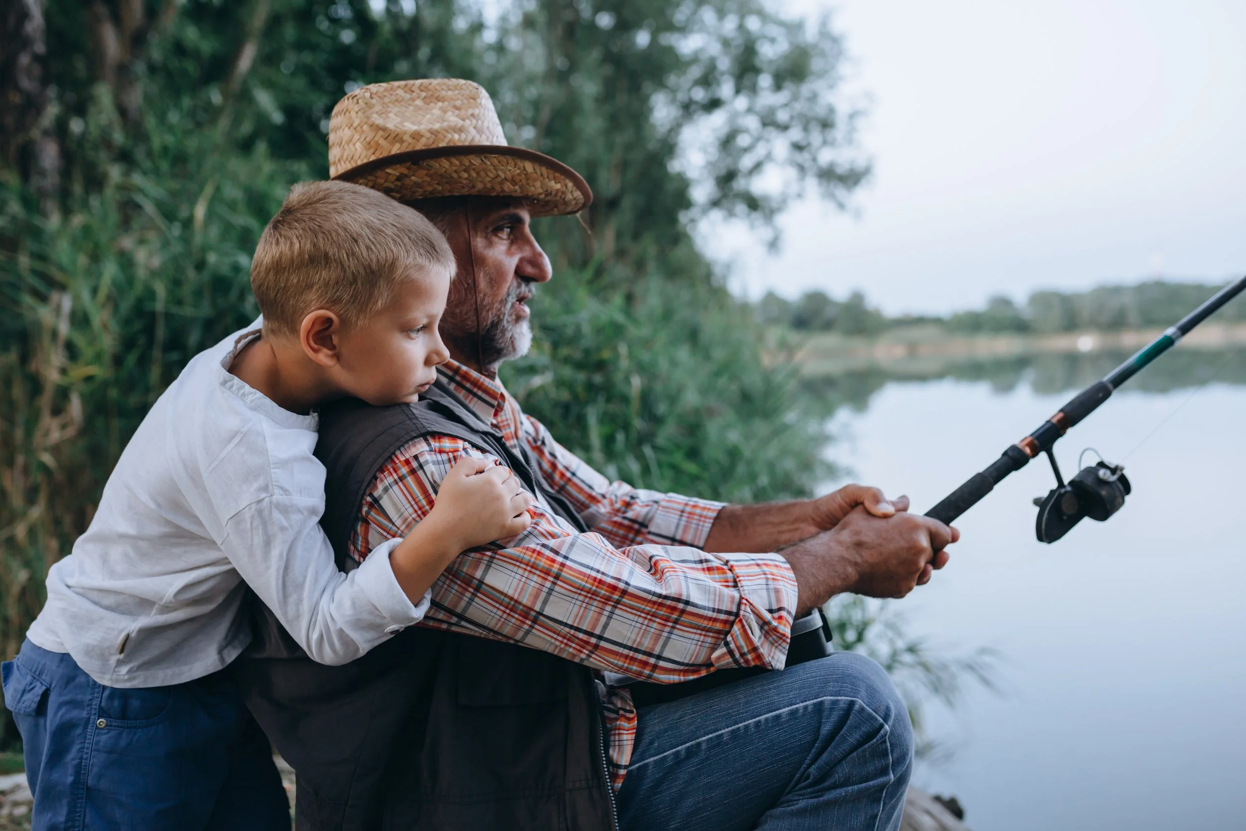 An elderly man fishing by a lake with a young boy embracing him from behind.