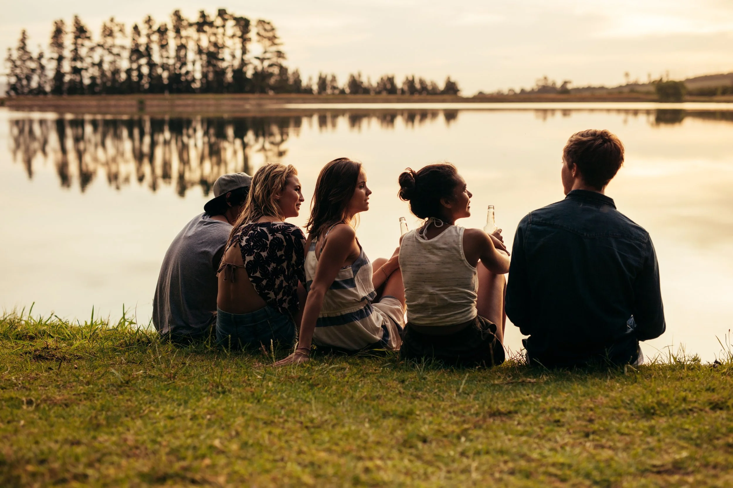 Five friends sitting on grass by a lake at sunset, chatting and holding bottles.