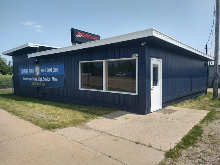 A blue building with a sign announcing the launch of a boat club, located on a sidewalk with grass nearby, and a Mercury sign visible in the background.