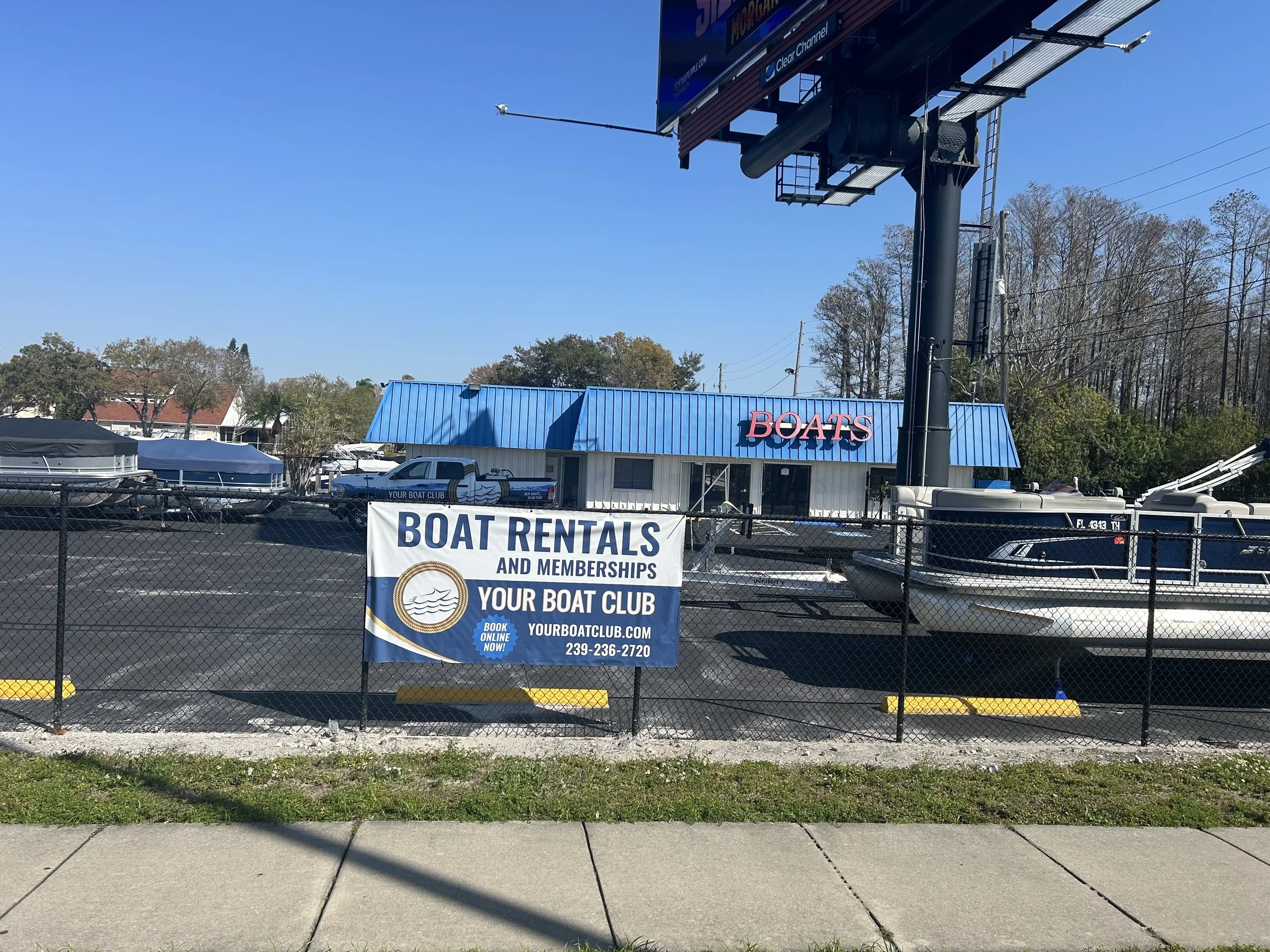 A boat rental business with a sign that reads 'BOAT RENTALS AND MEMBERSHIPS YOUR BOAT CLUB', located behind a black chain-link fence. There are boats parked in the lot and a building with a blue roof and a large sign that says 'BOATS'.