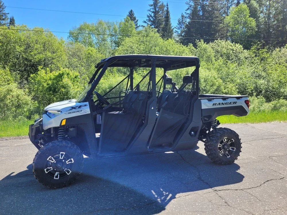 A side view of a black and white Polaris Ranger off-road utility vehicle parked on an asphalt surface with green trees and blue sky in the background.
