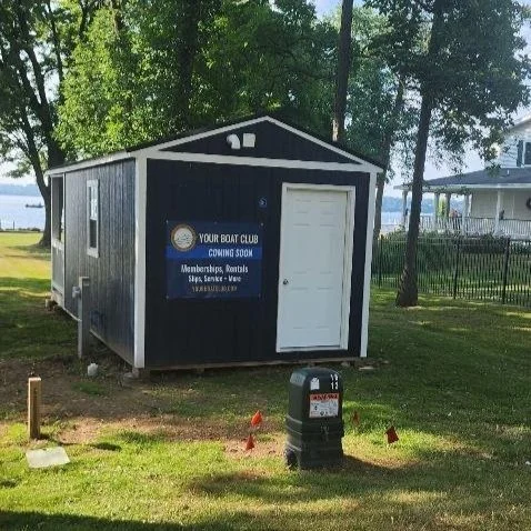 Small black and white building with a sign that reads "Your Boat Club Coming Soon," located in a grassy area with trees and a fence nearby.