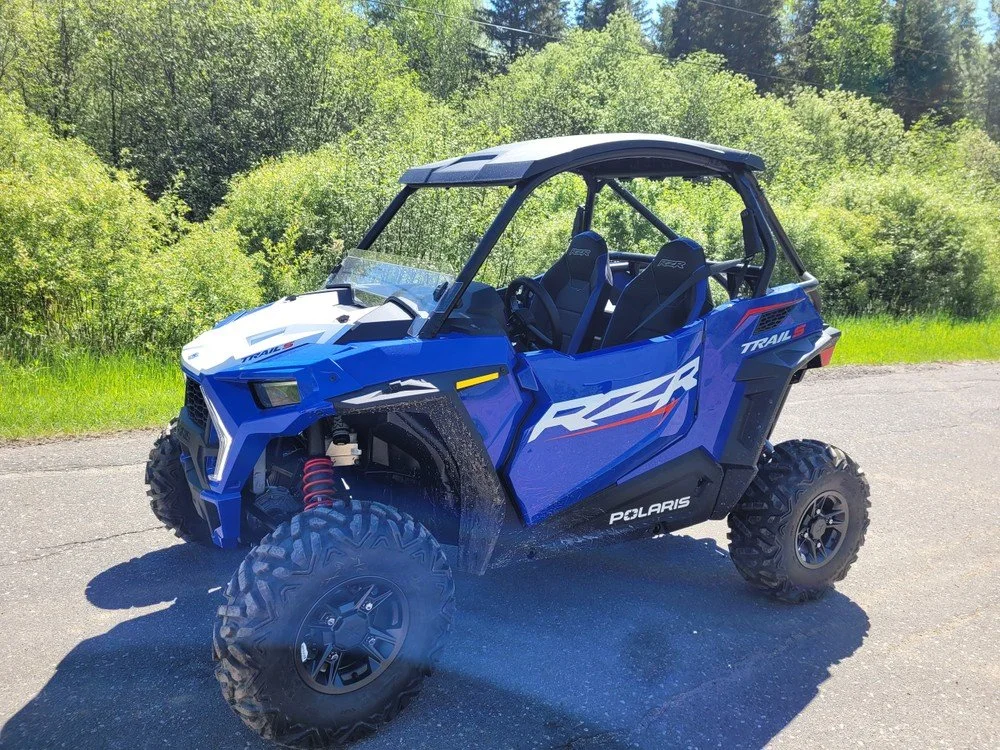 Blue Polaris RZR off-road vehicle parked on a paved surface with green trees and bushes in the background.