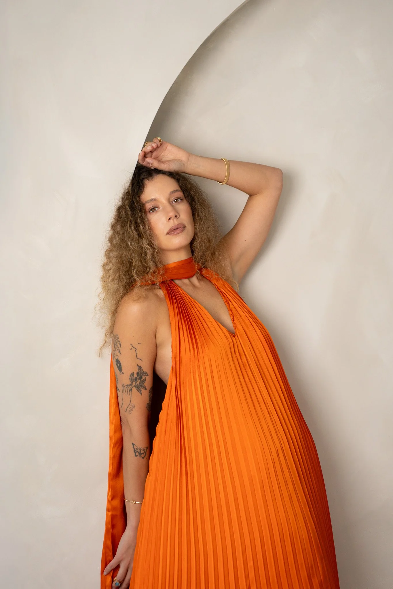 A woman with curly hair posing against a white wall, wearing an orange pleated dress and gold jewelry, with tattoos on her left arm.