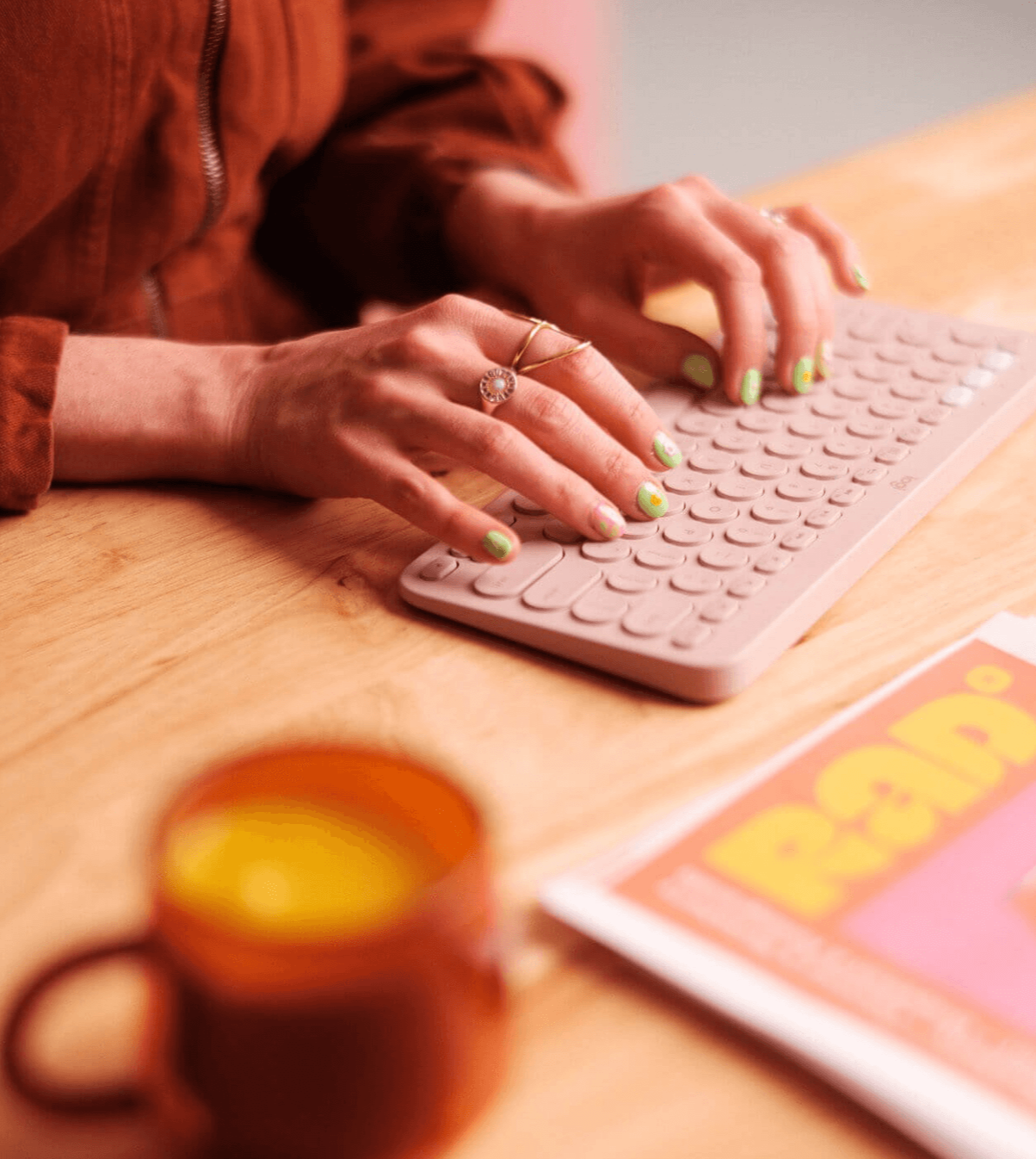 Close-up of a person using a pink keyboard, wearing rings and with green and yellow nail polish, on a wooden desk with a colorful magazine and a blurry orange mug in the foreground.
