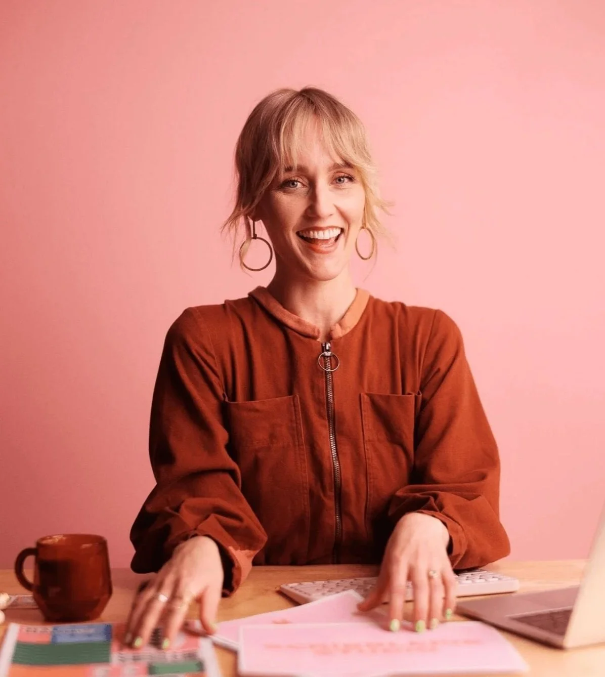 A woman with short blonde hair and hoop earrings, smiling and sitting at a desk against a pink wall. She is wearing a rust-colored jacket with a zipper and appears to be working or studying, with papers, a keyboard, and a mug on the desk.