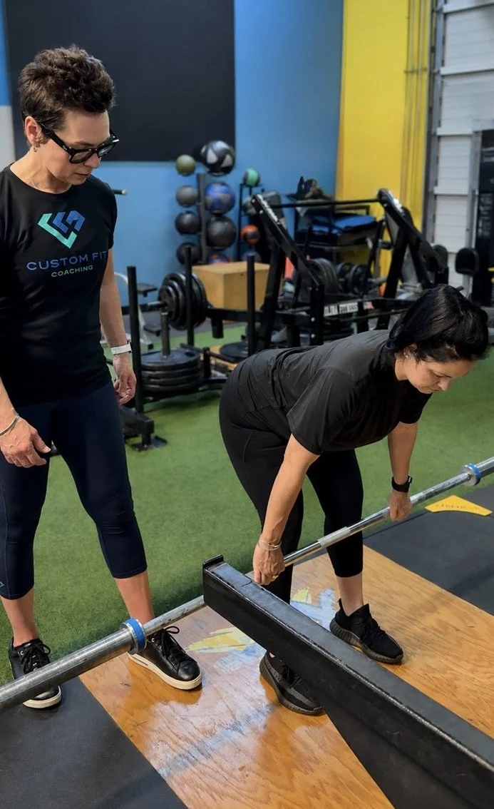 Woman performing a deadlift with a barbell while a coach oversees at a gym with weightlifting equipment in the background.