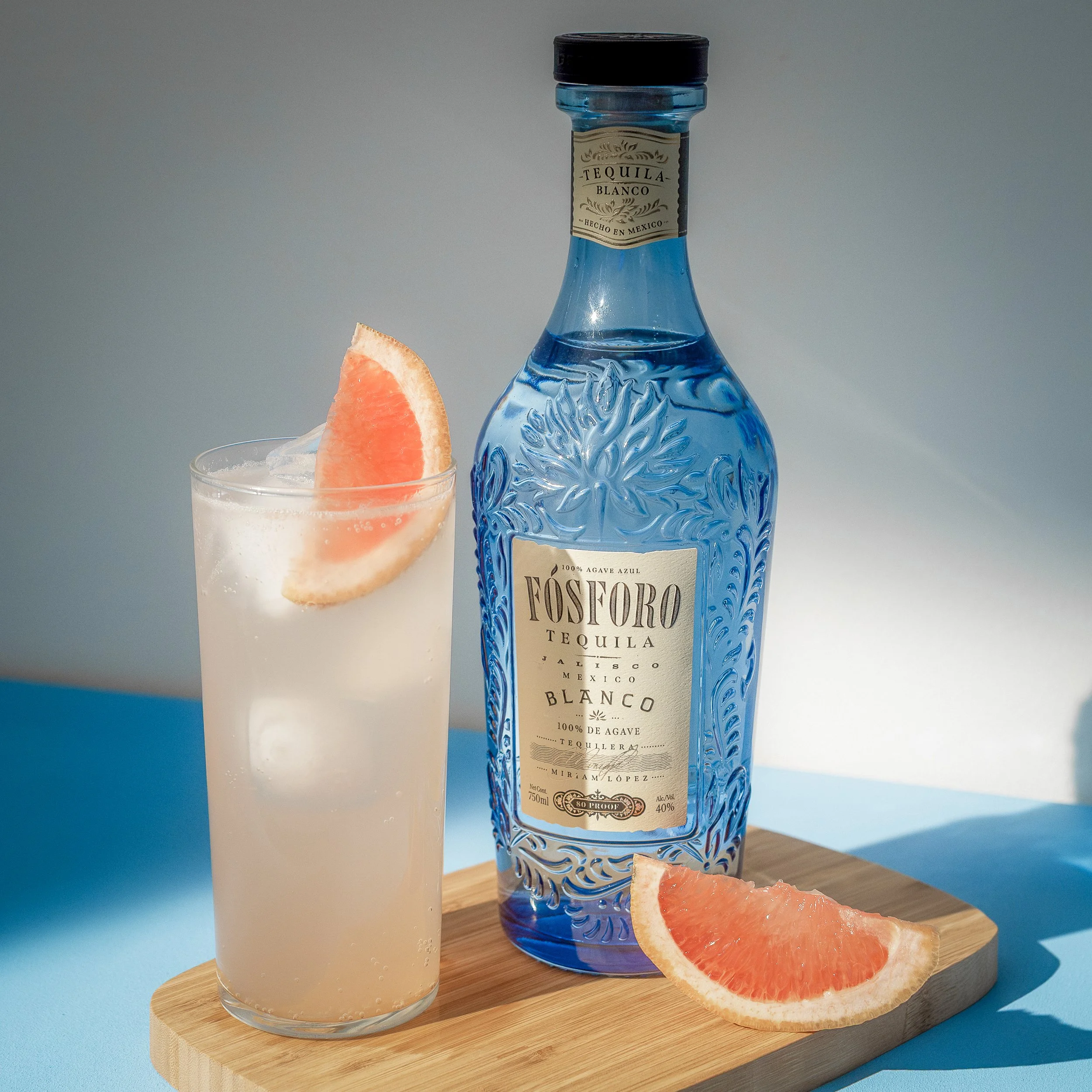A paloma in a highball glass garnished with a grapefruit wedge on the rim, a bottle of Fósforo Tequila, and a grapefruit slice on a wooden board with a pale blue and gray background.