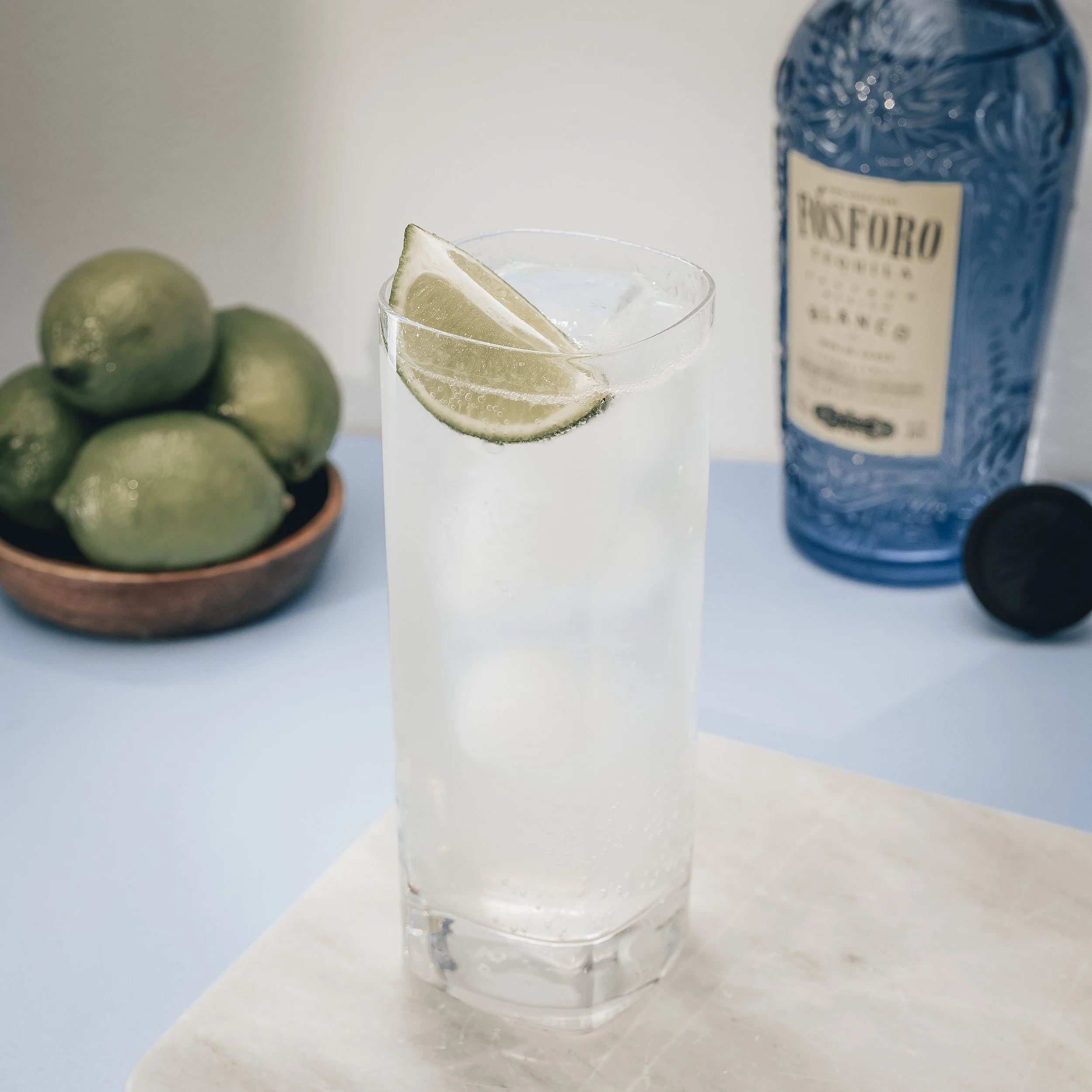 A ranch water cocktail in a highball glass garnished with a lime wedge, with a bowl of limes and a bottle of Fósforo Tequila Blanco in the background.