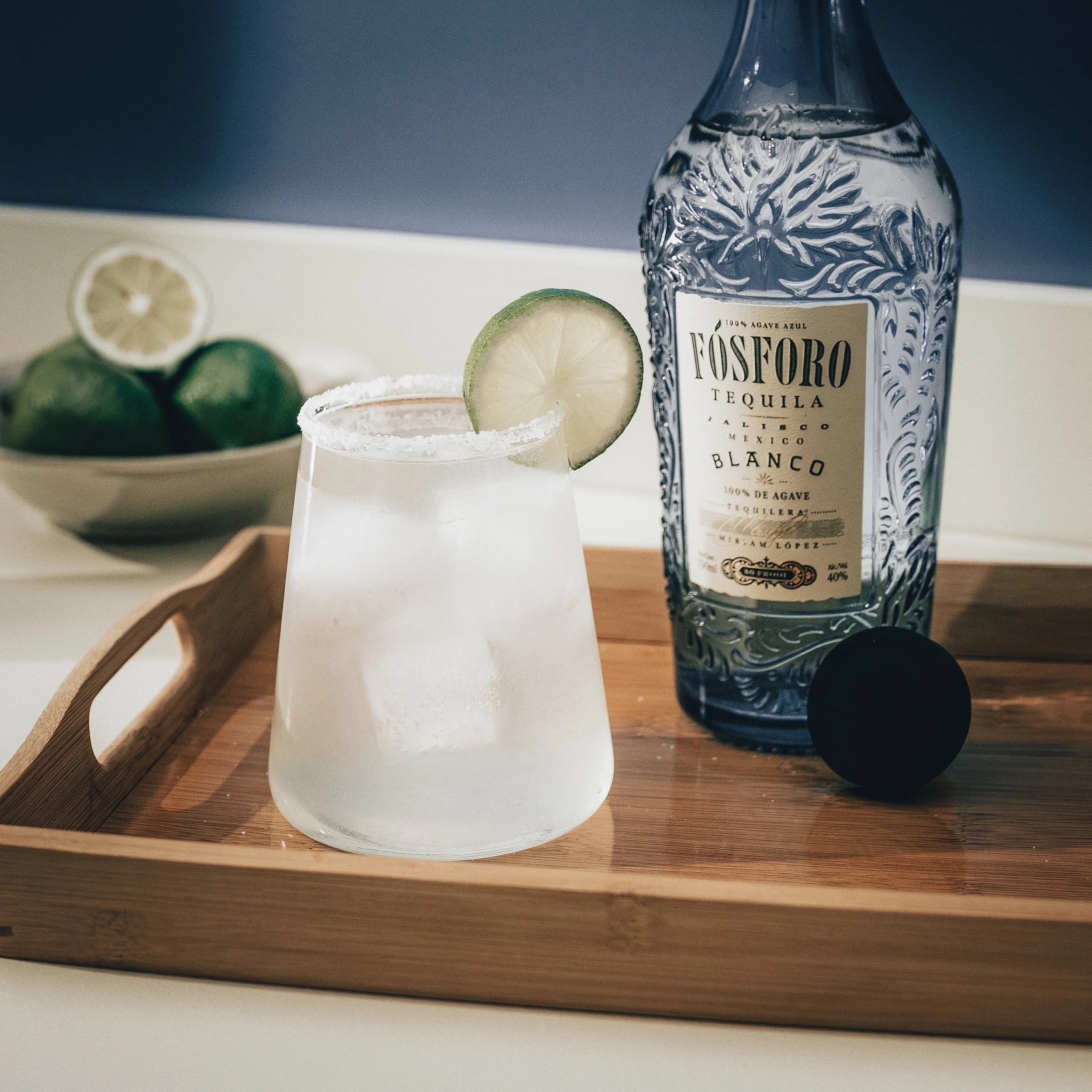 A margarita in a rocks glass with a salted rim and lime garnish, a bottle of Fósforo Tequila, and a bowl of limes on a wooden tray.