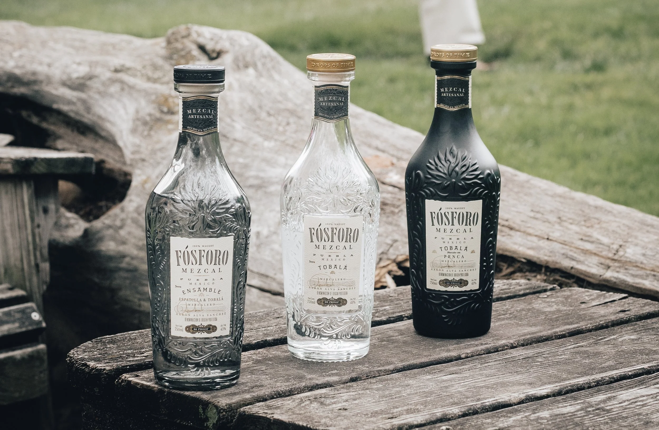 Three bottles of Fósforo Mezcal on a wooden table outdoors, with one Tobalá, one Ensamble, and one Penca, all showcasing intricate embossed designs and labels.