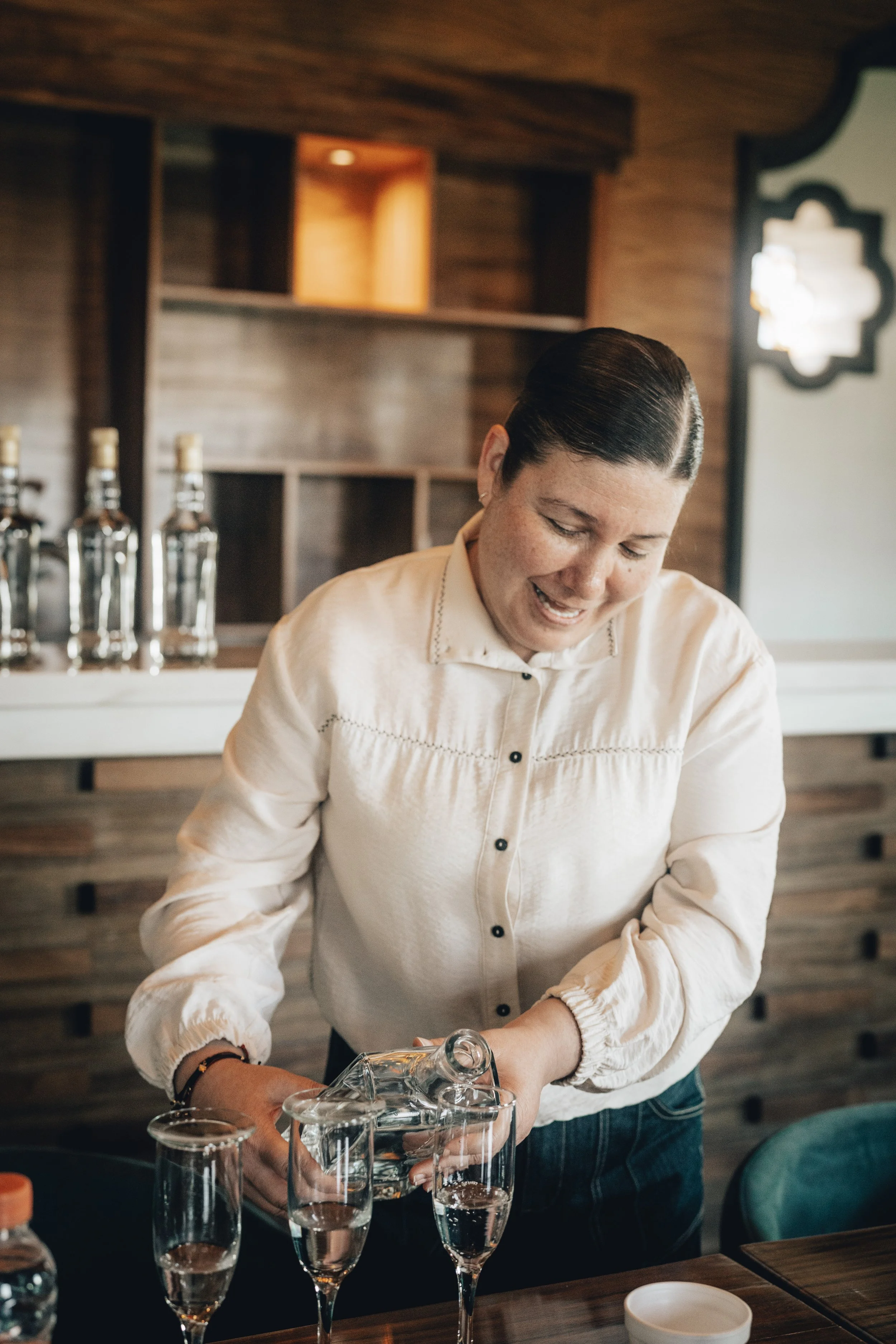 Tequilara Miriam Lopez pouring a Fósforo Tequila into champagne glasses in a tasting room with wooden decor.