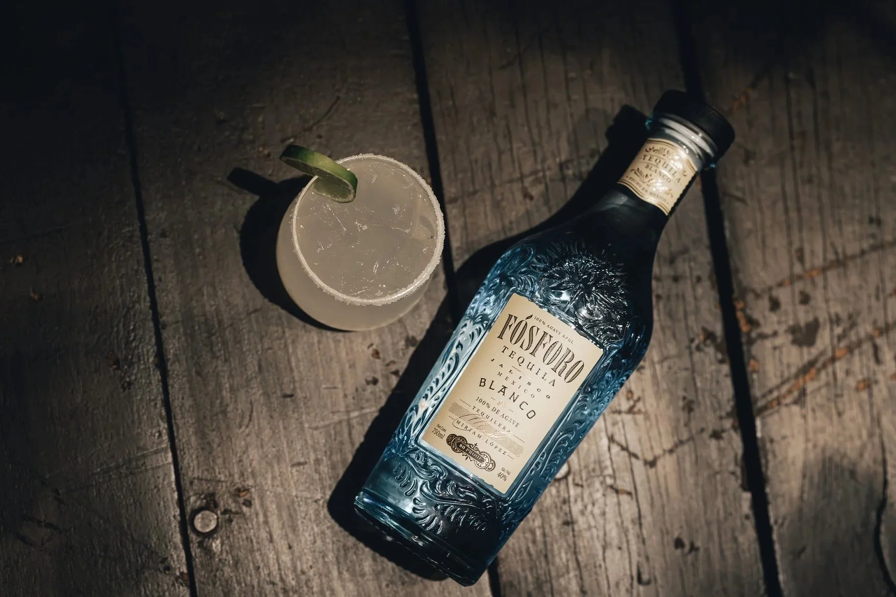 A bottle of Fósforo tequila lying on a wooden surface next to a glass of margarita with a salted rim and a lime wedge.