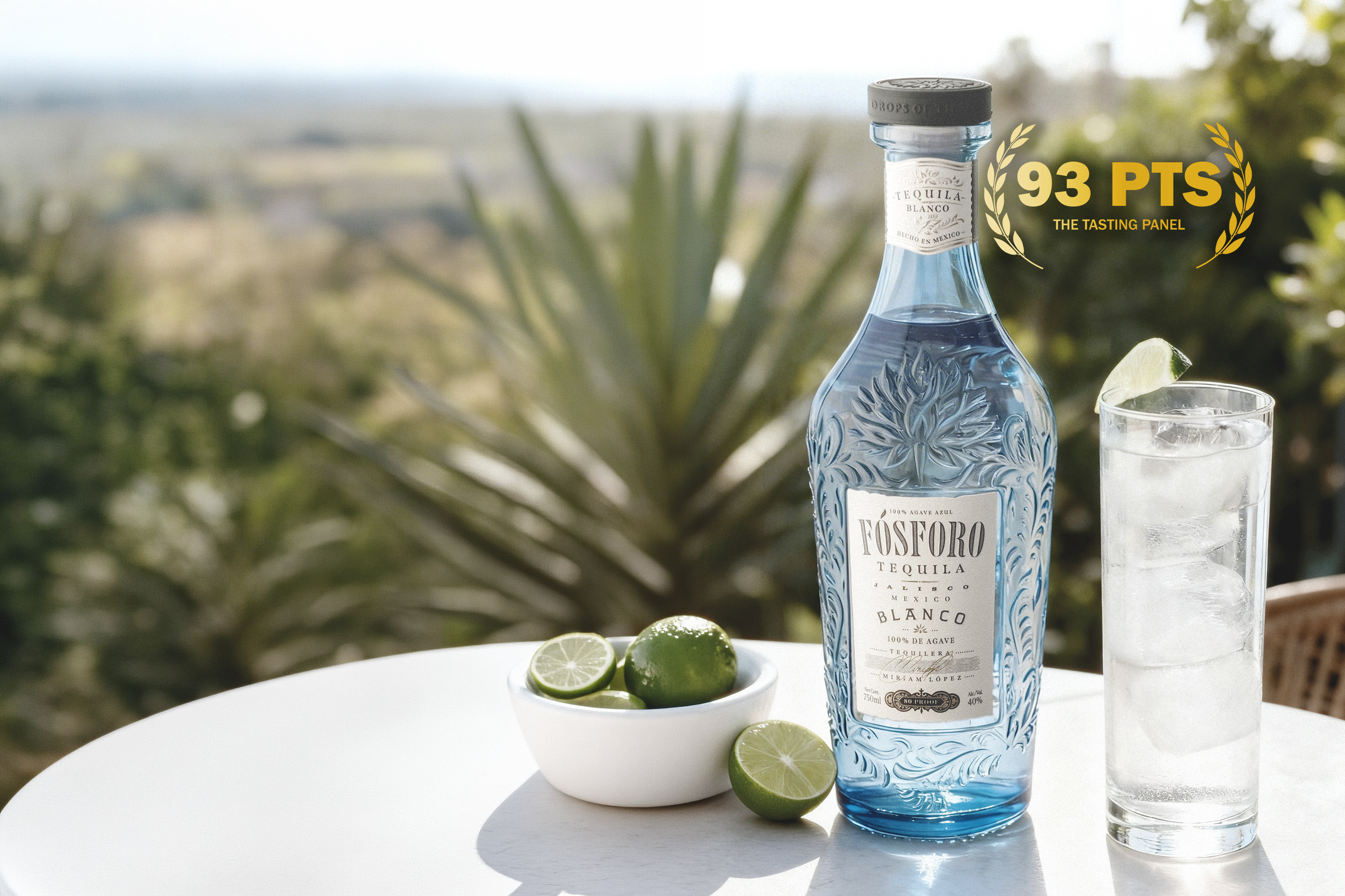Bottle of Fósforo tequila, a ranch water in a highball glass with ice and lime wedge, and a bowl of lime halves on a white table outdoors with a blurred background of greenery.
