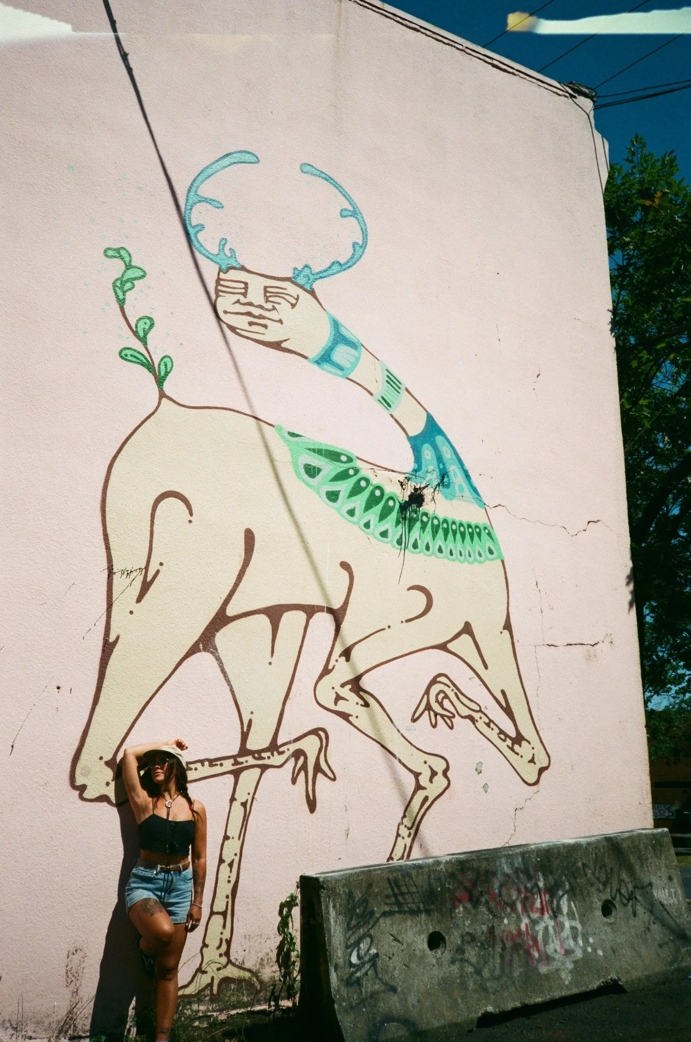 Mural art on a wall depicting a surreal creature with elongated limbs and foliage designs, and a person standing at the base in casual attire.
