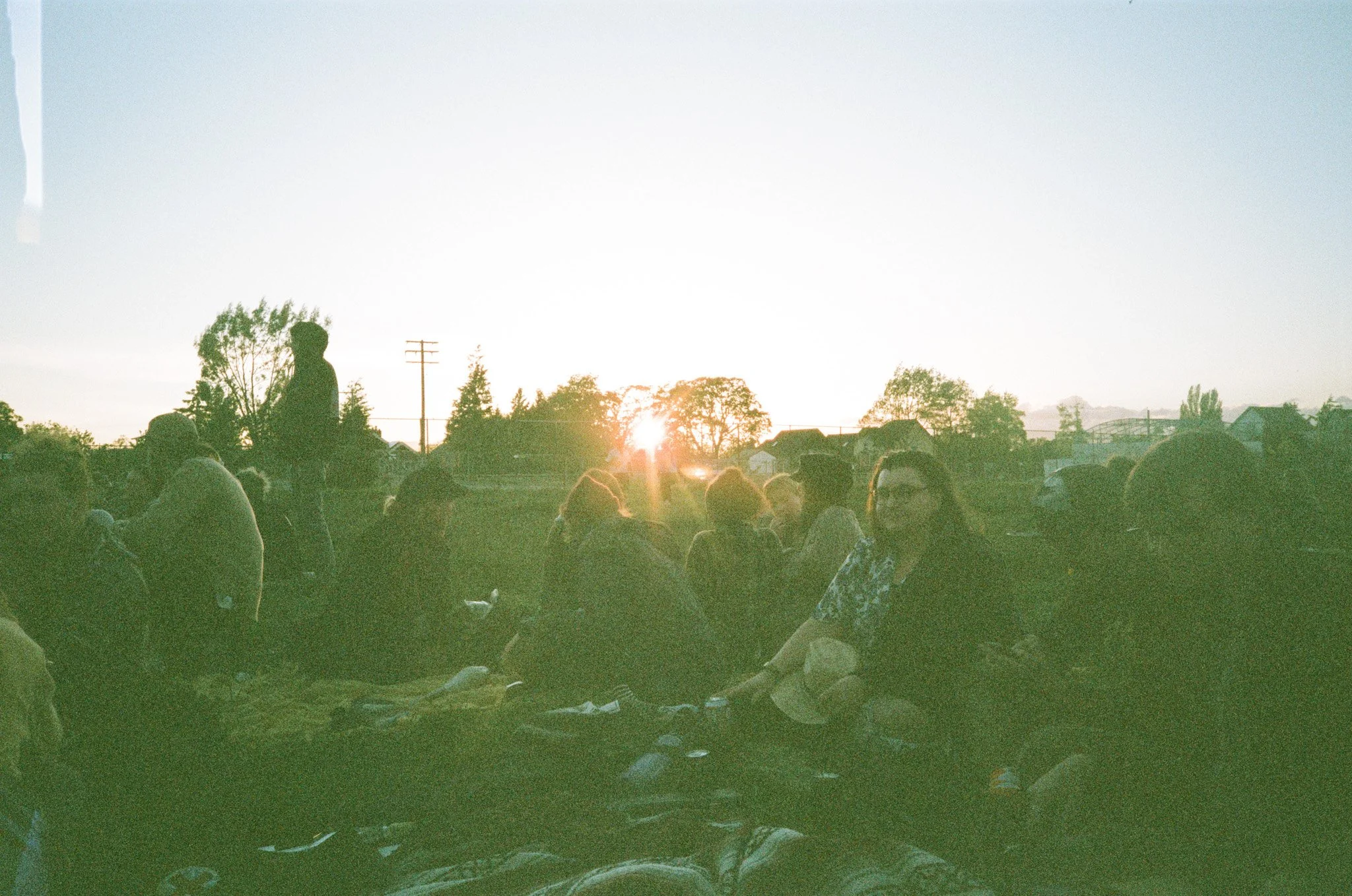 Group of people sitting outdoors during sunset, silhouetted against the sky, with trees and power lines in the background.