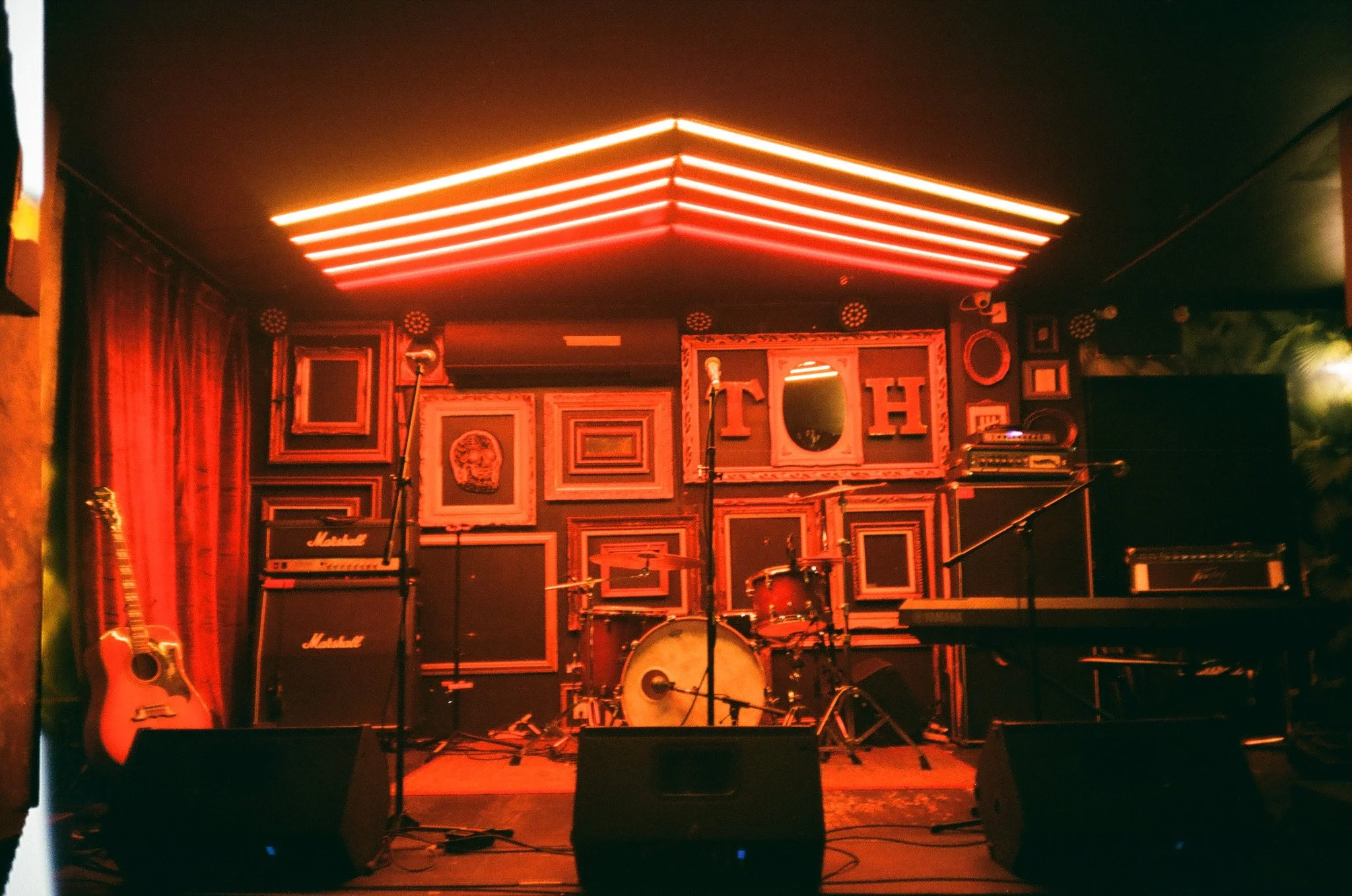 Empty music stage with red lighting, featuring a drum kit, microphone stands, a guitar, amplifiers, and a wall decorated with framed art pieces. TurboHaus, Montreal