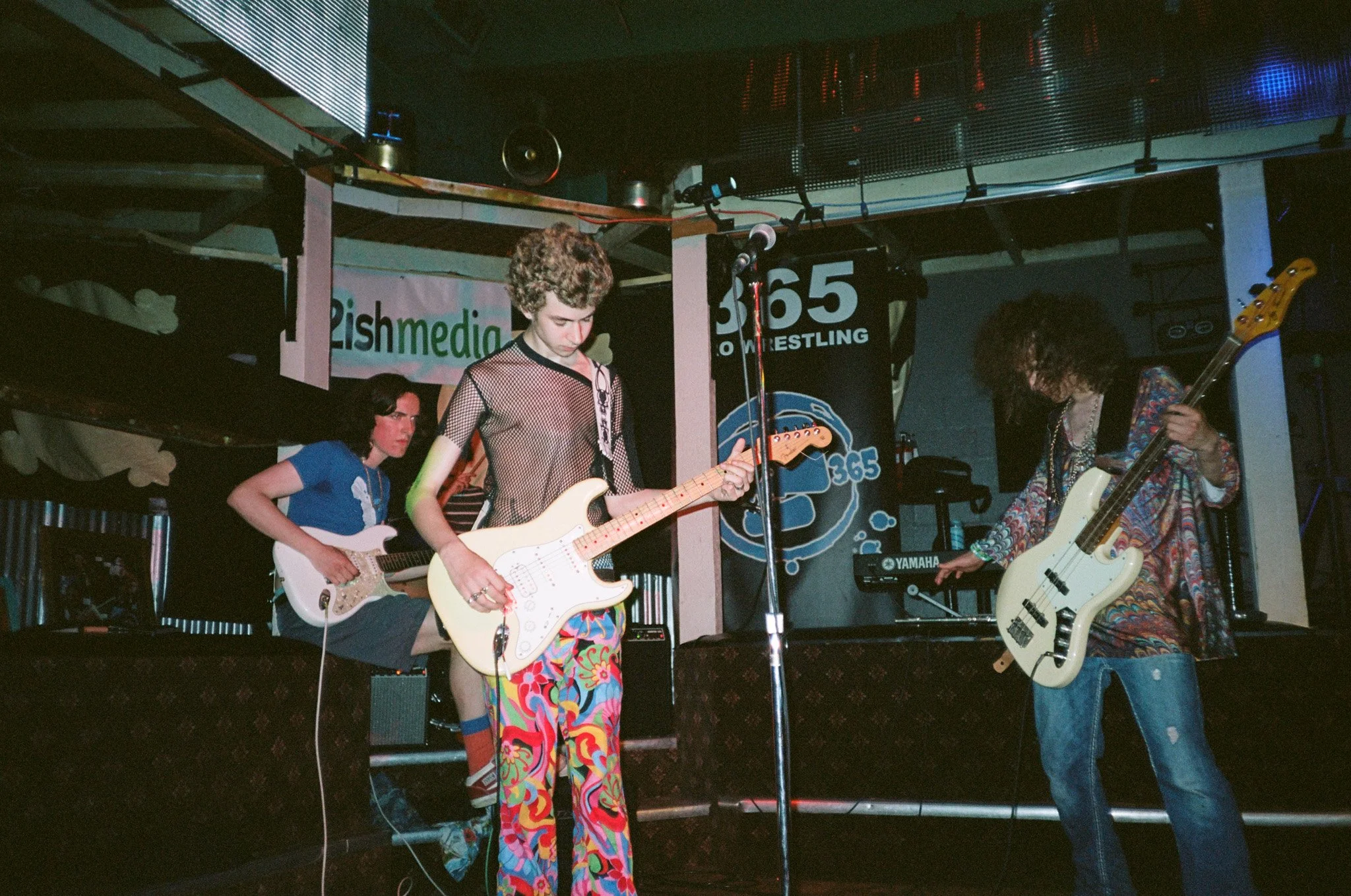 Band performing on stage with electric guitars and bass, vibrant clothing, music venue with banners in background.