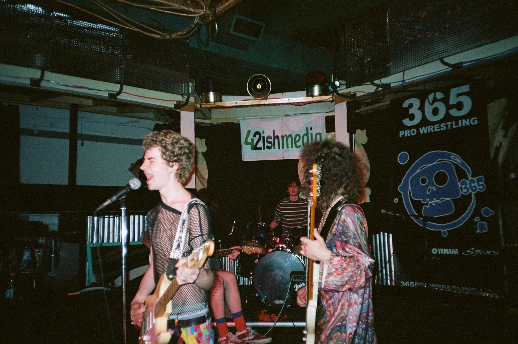 Band performing on stage with guitars and drums, backdrop shows 42ishmedia and 365 Pro Wrestling logos, indoor setting.