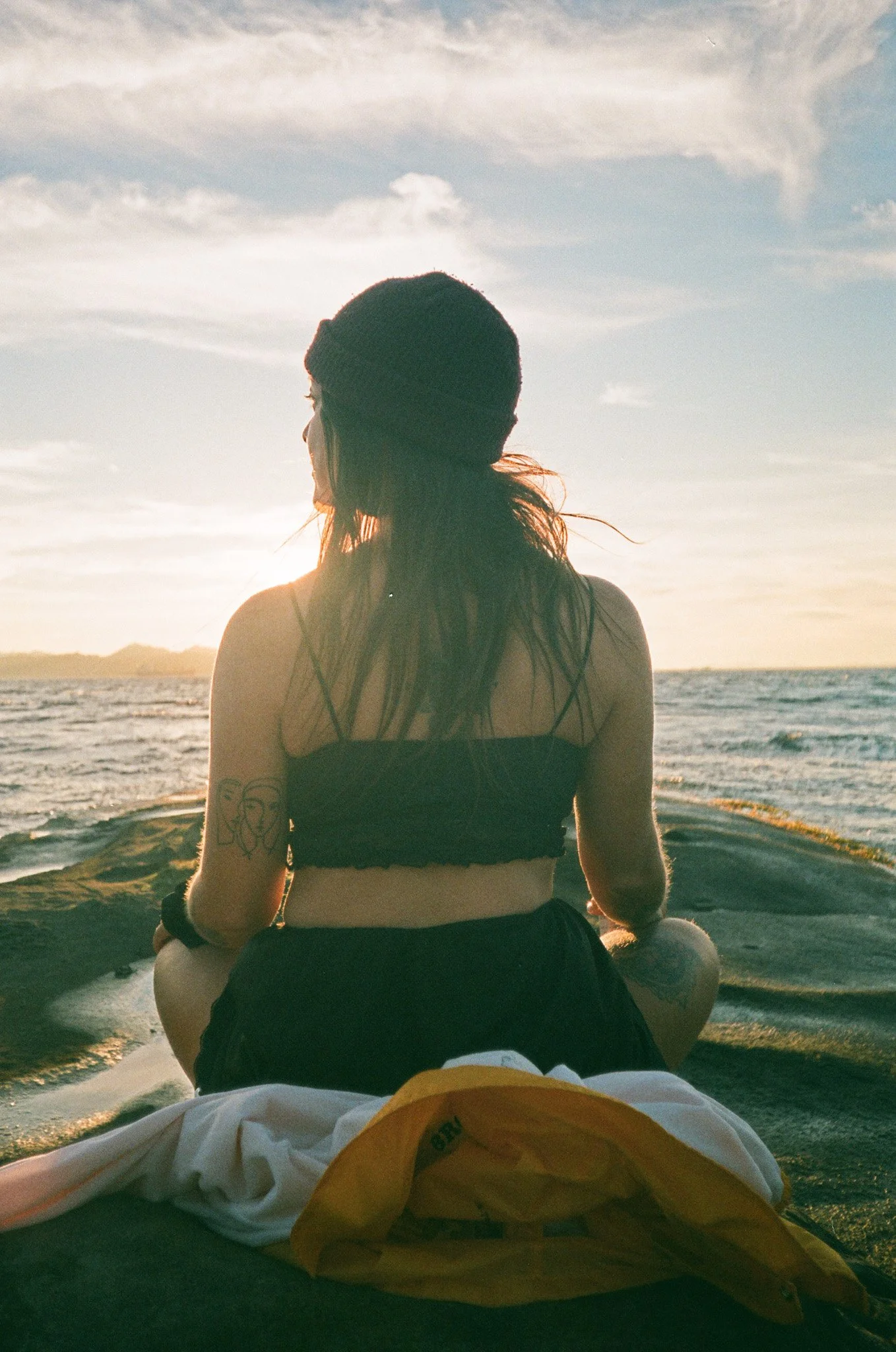 Person sitting on a rock by the sea at sunset, wearing a beanie and a top, with a tattoo on the arm.