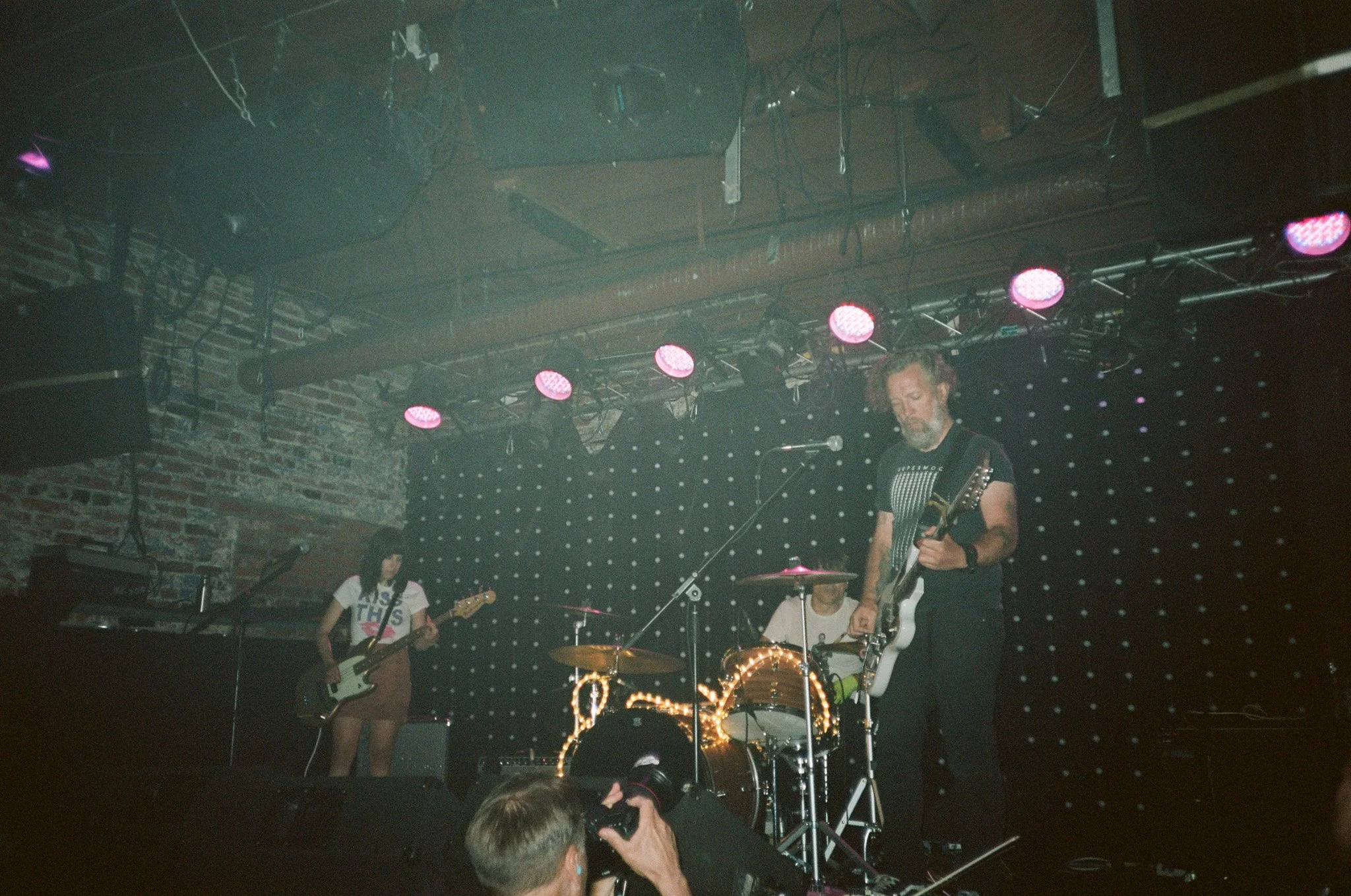 Live band performing on dimly lit stage with guitar, bass, and drums, brick wall backdrop and purple spotlights.