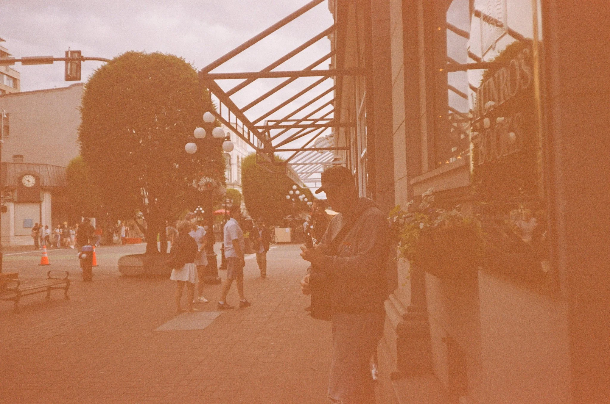Street scene with people walking and a musician playing guitar near a bookstore; trees and vintage street lamps line the sidewalk.