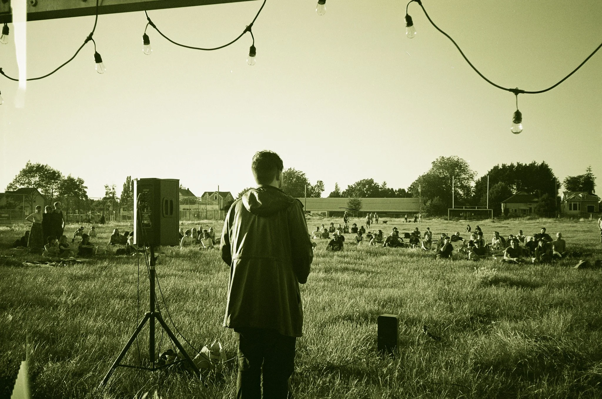 Person speaking to an outdoor seated audience in a field with string lights overhead.