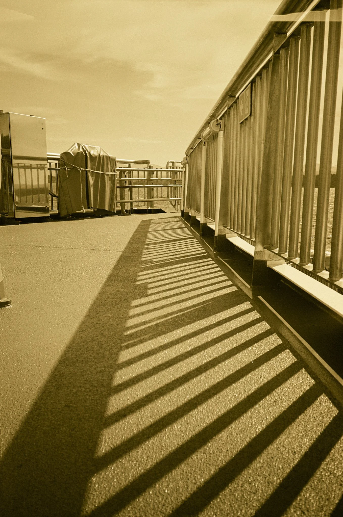 Sepia-toned photo of a rooftop with metal railings, shadows cast on the ground, and covered equipment in the background under a clear sky.