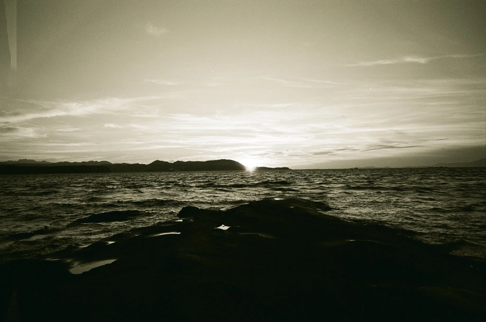 Monochrome coastal sunset with rocky shore, ocean waves, and distant hills.