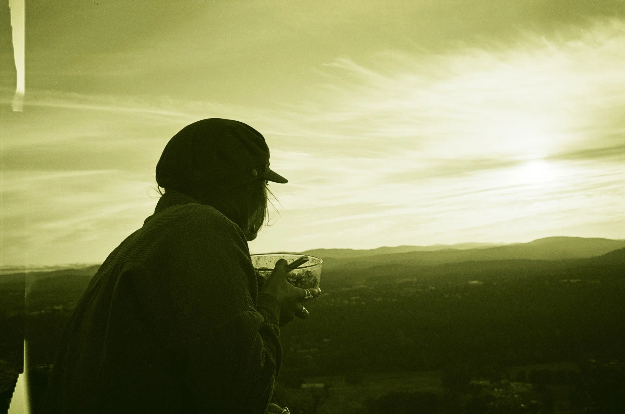 Silhouette of a person wearing a hat, holding a bowl while looking at a scenic sunset view.
