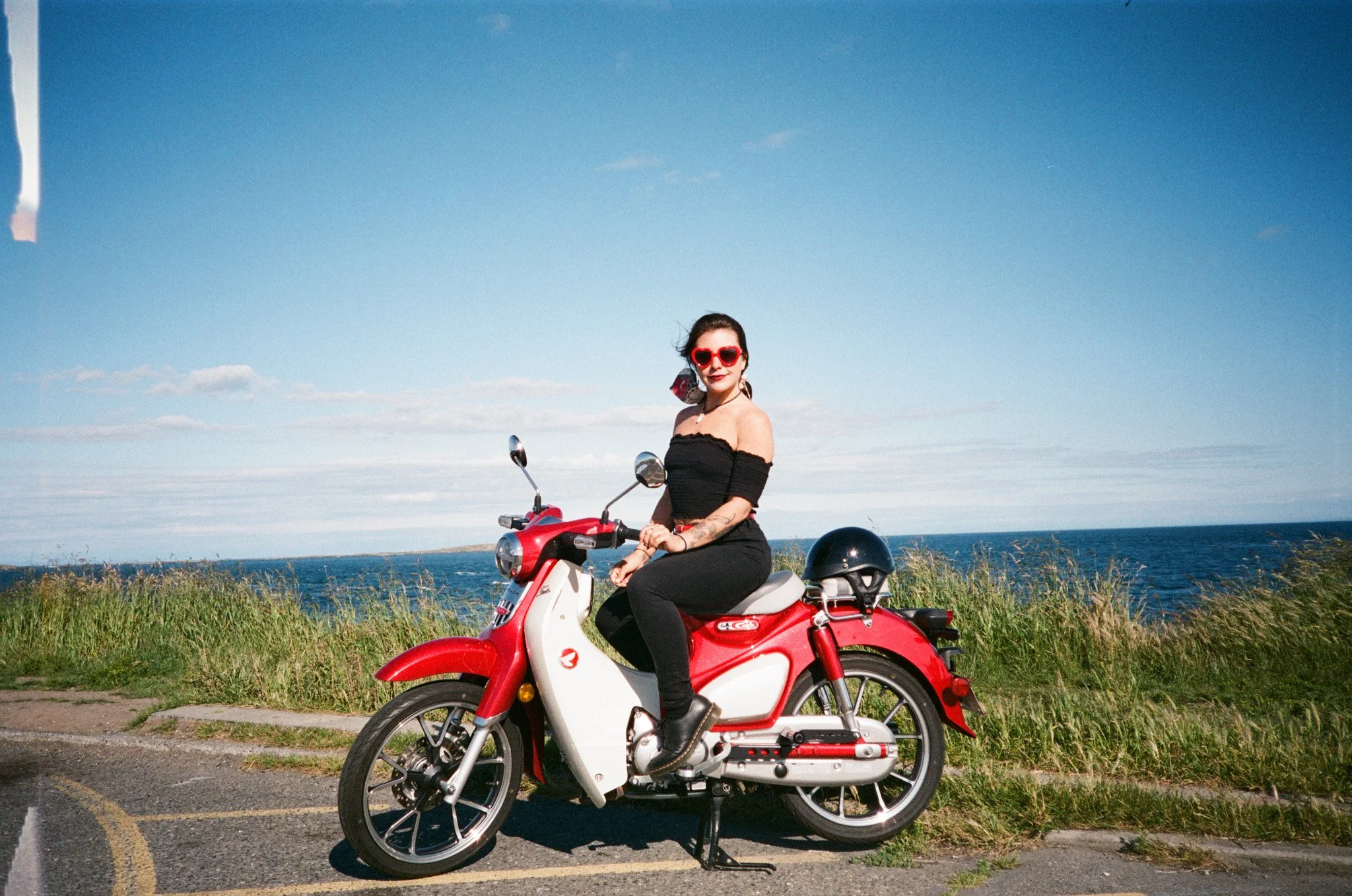 Person posing on a red motorcycle near the ocean with grass and blue sky.