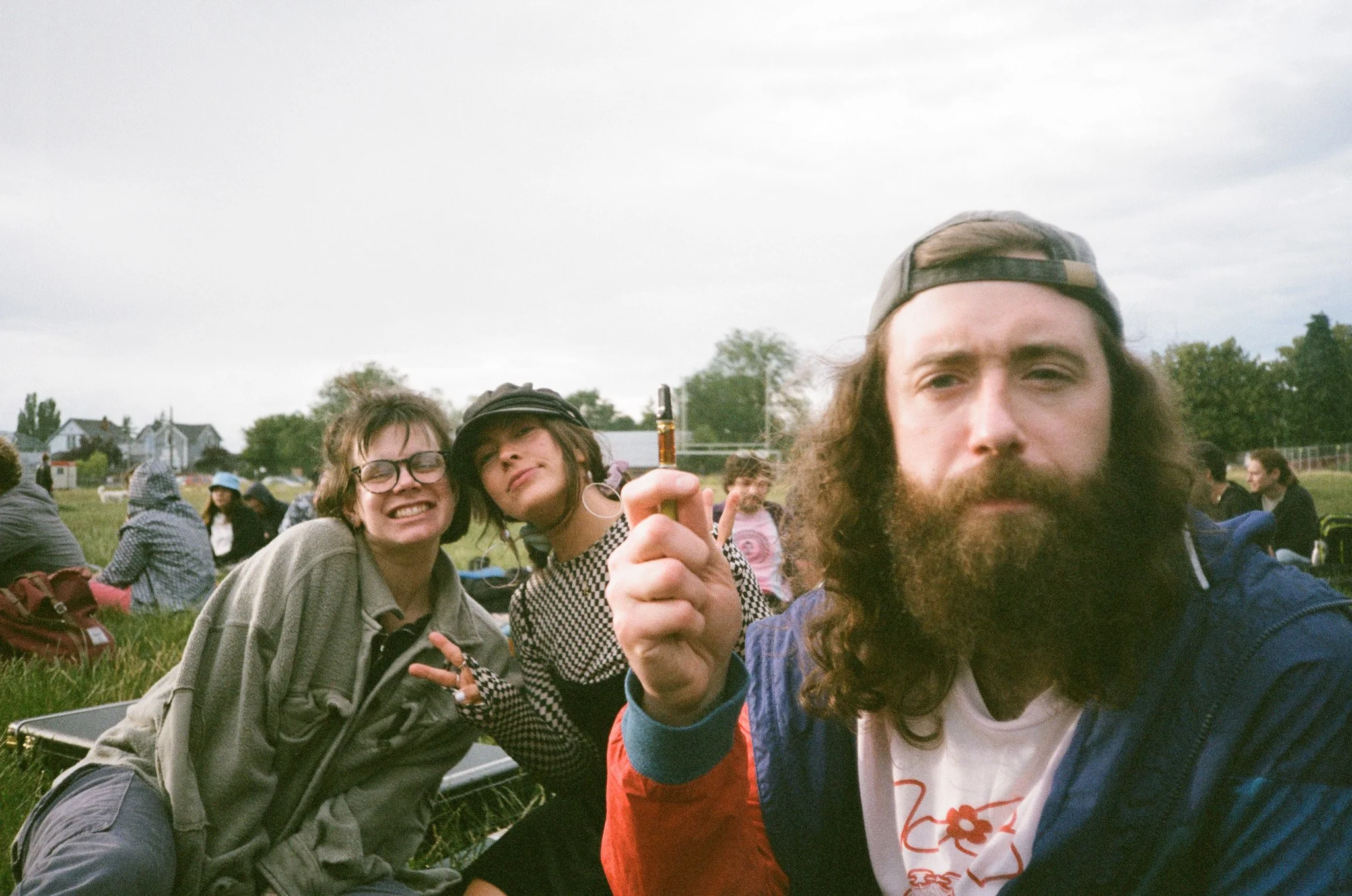 Three people sitting outdoors on grass, one holding a vape pen, with others in the background.
