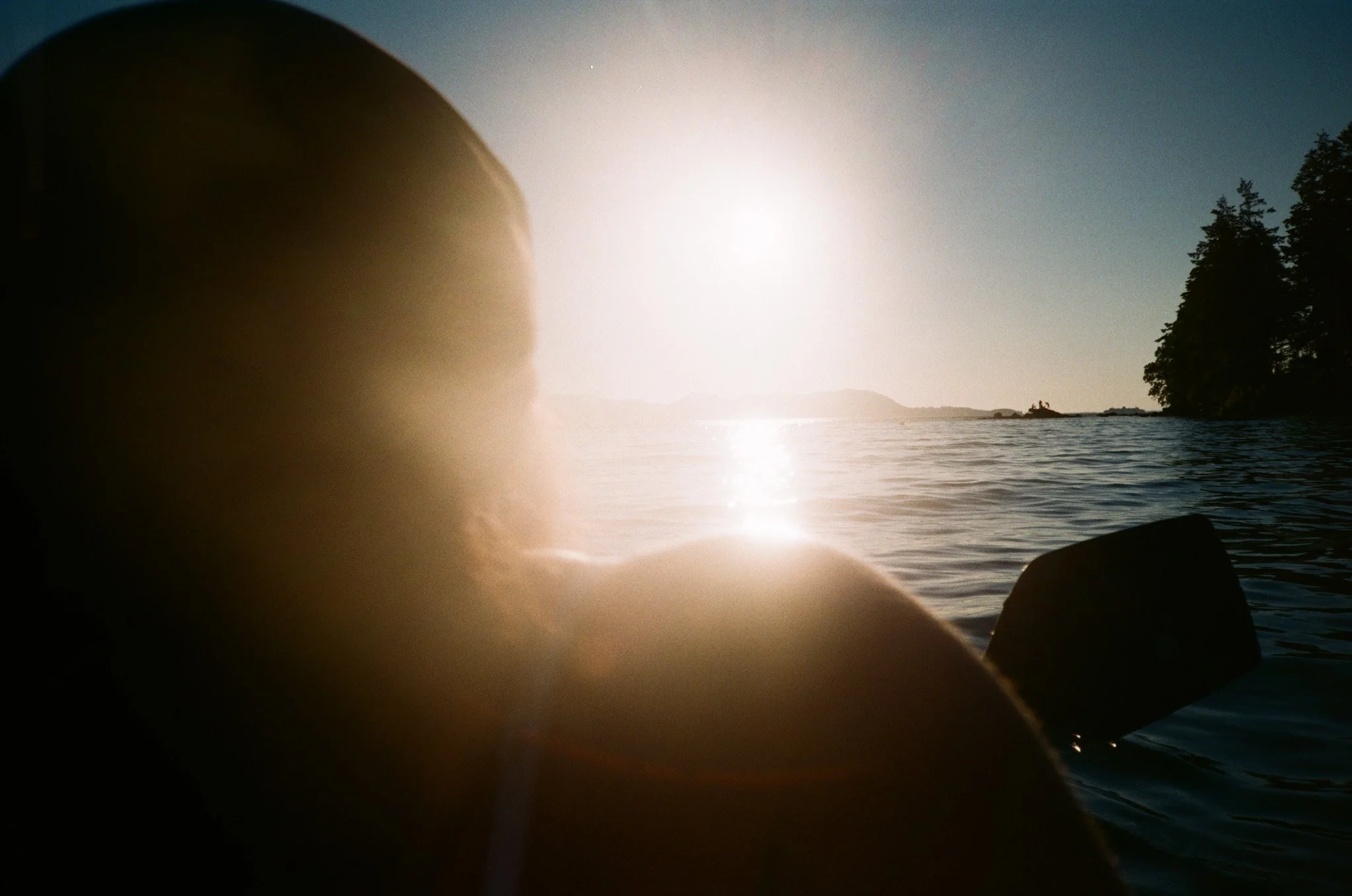 Silhouette of person kayaking on a calm lake at sunset with distant trees and islands visible.