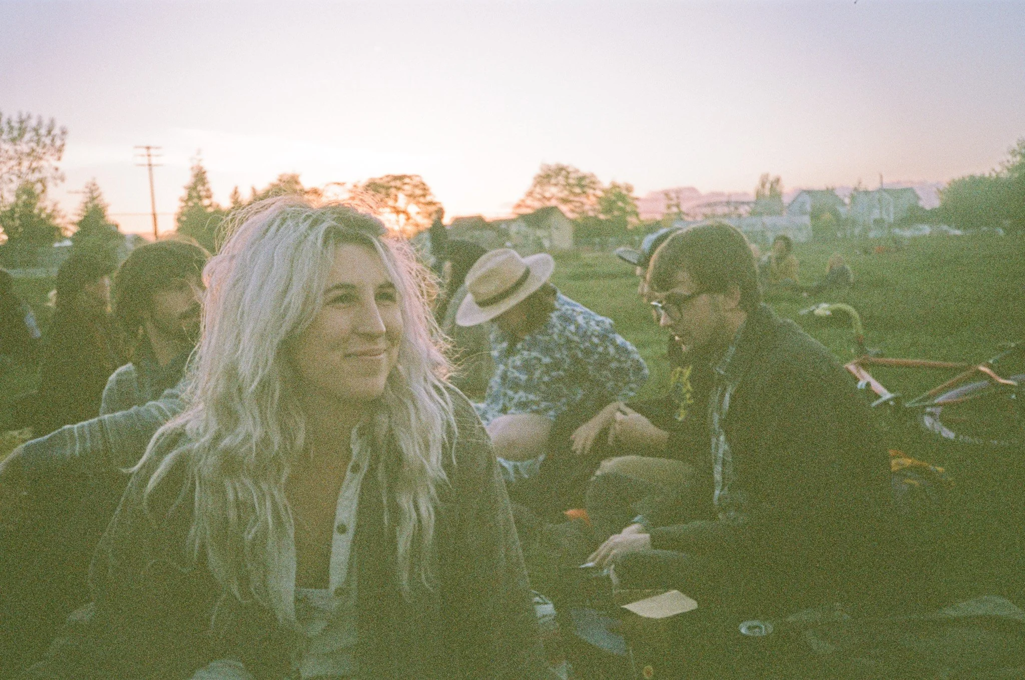 Group of people sitting outdoors at sunset, with one person in focus smiling, wearing a casual outfit. The background includes trees and a soft evening sky.