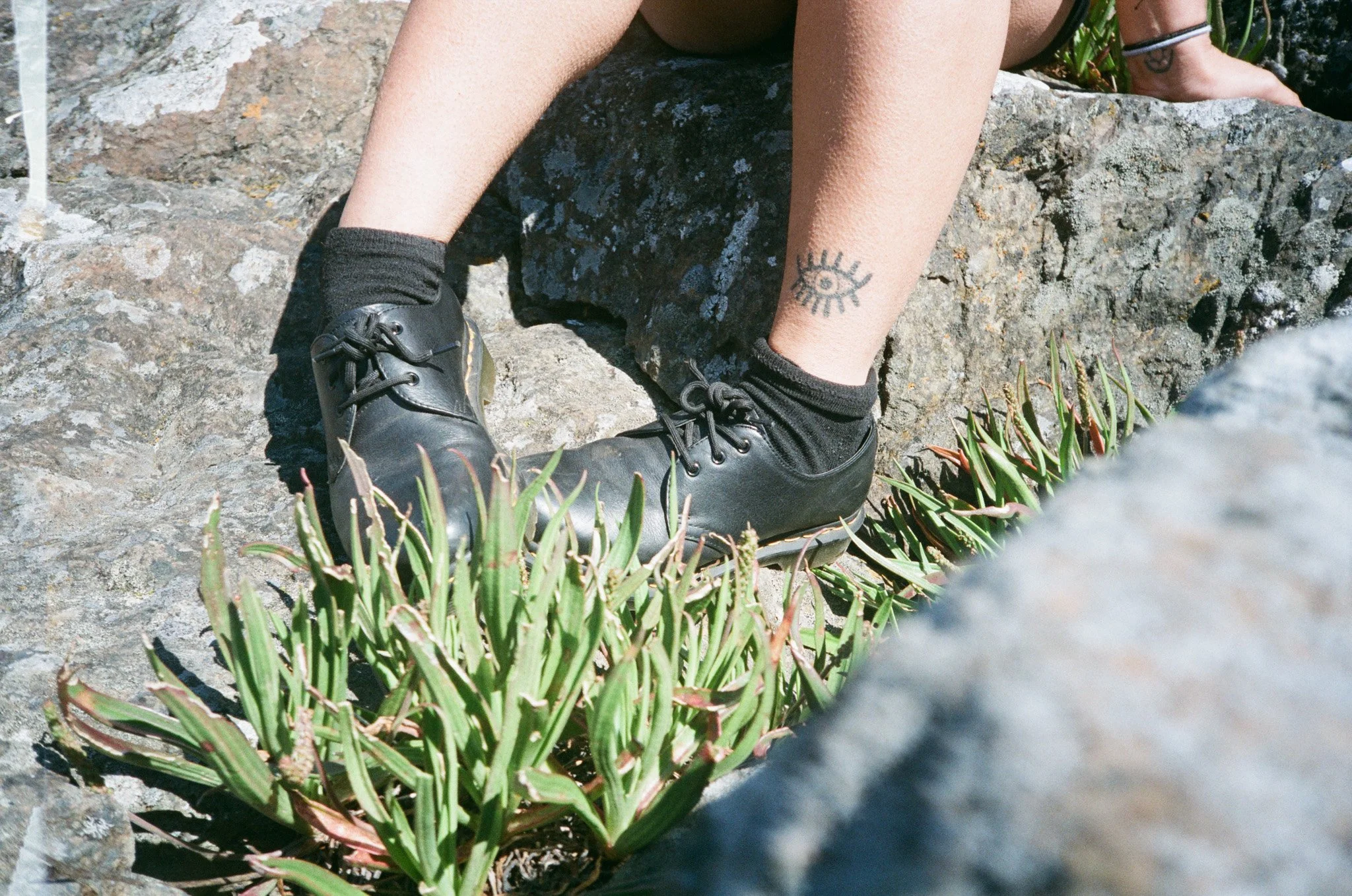 Person wearing black boots sitting on rocks with tattoo of an eye on their leg