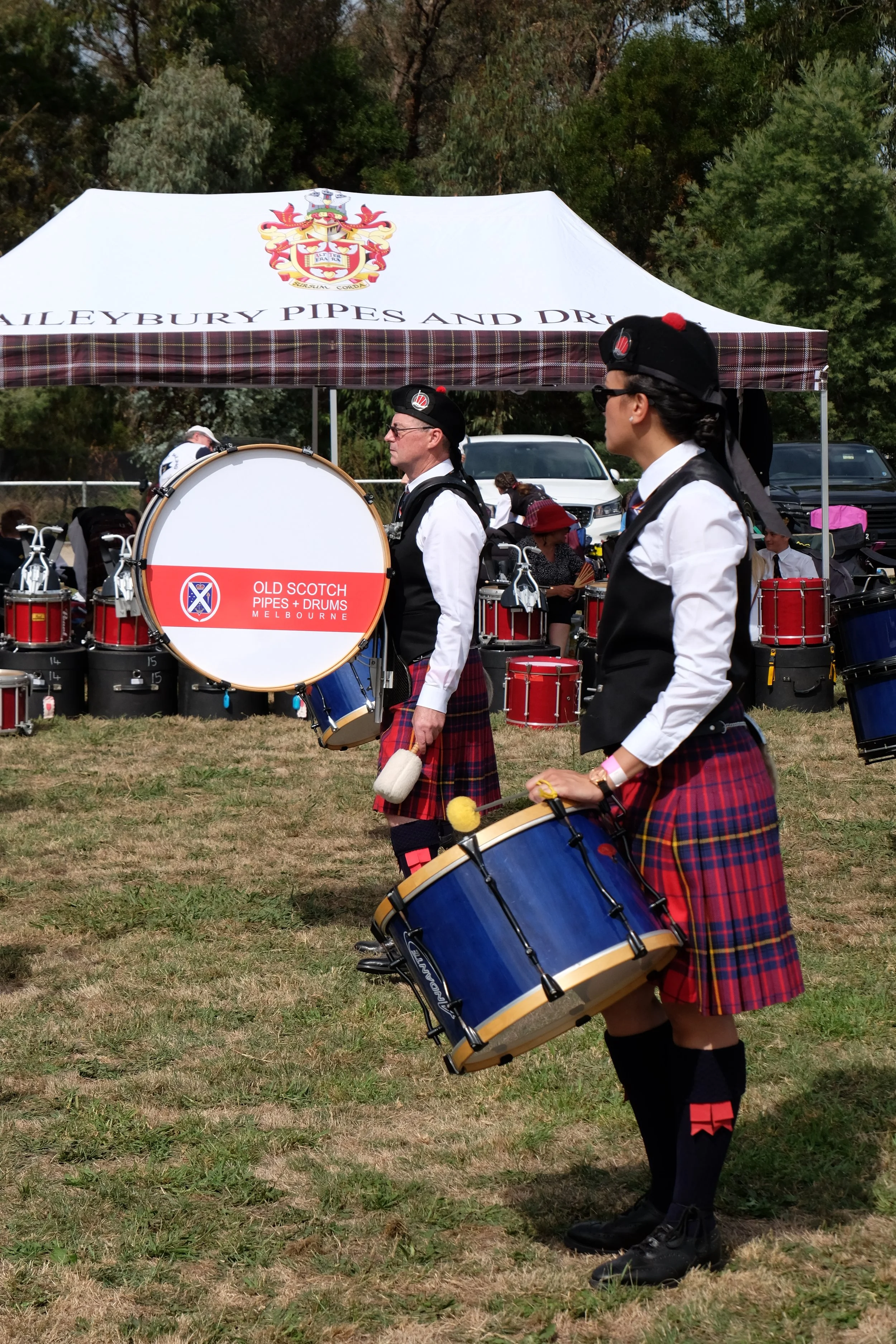 Two drummers in traditional Scottish attire, including kilts, performing at an outdoor event with a tent and other drums in the background.