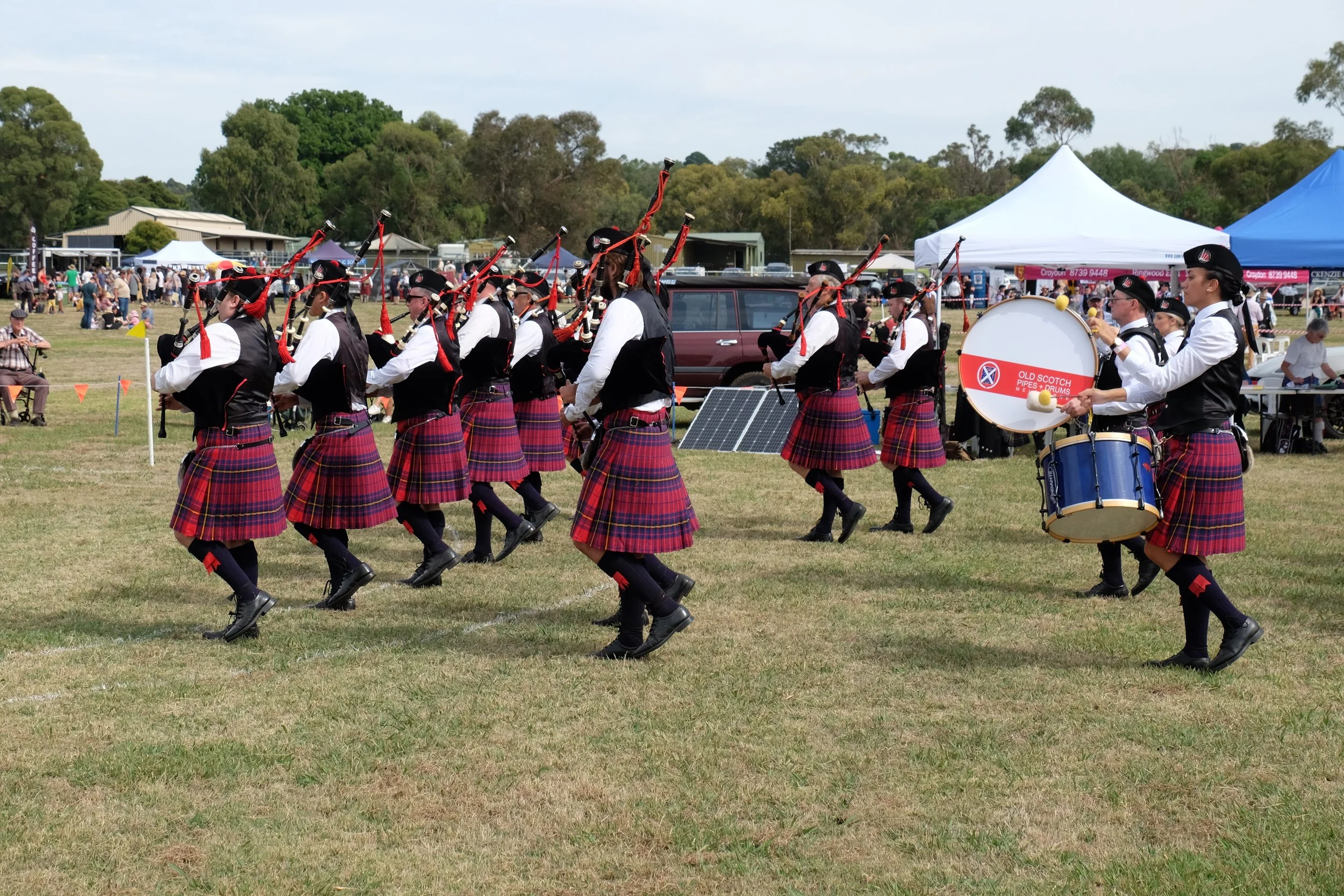 A group of musicians dressed in traditional Scottish uniforms playing bagpipes and drums at an outdoor event, with tents and people in the background.
