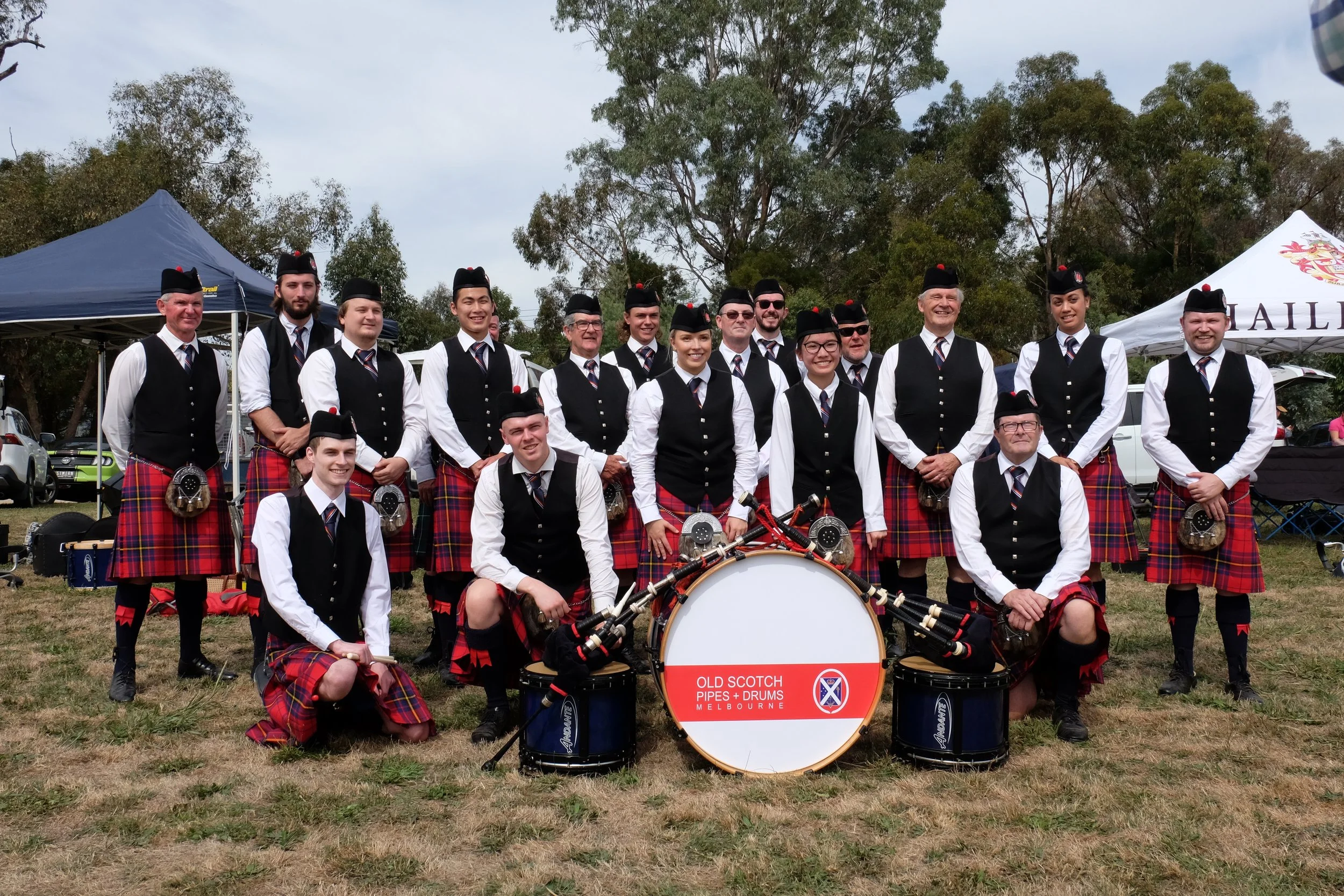 A group of people in traditional Scottish attire, including kilts, vests, and hats, standing outdoors on a grassy field with trees and tents in the background. They appear to be part of a pipe band, with drums and pipes visible, and a sign on the dru
