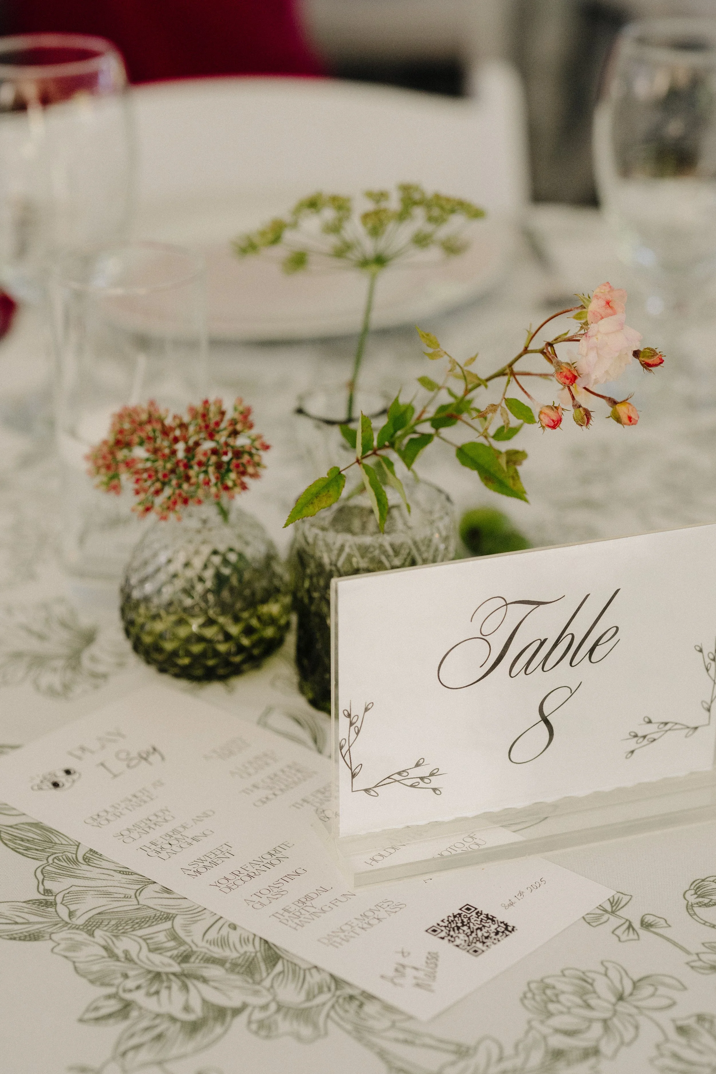 Wedding table setting with a table number card, floral decorations, and printed wedding program on a floral-themed tablecloth.