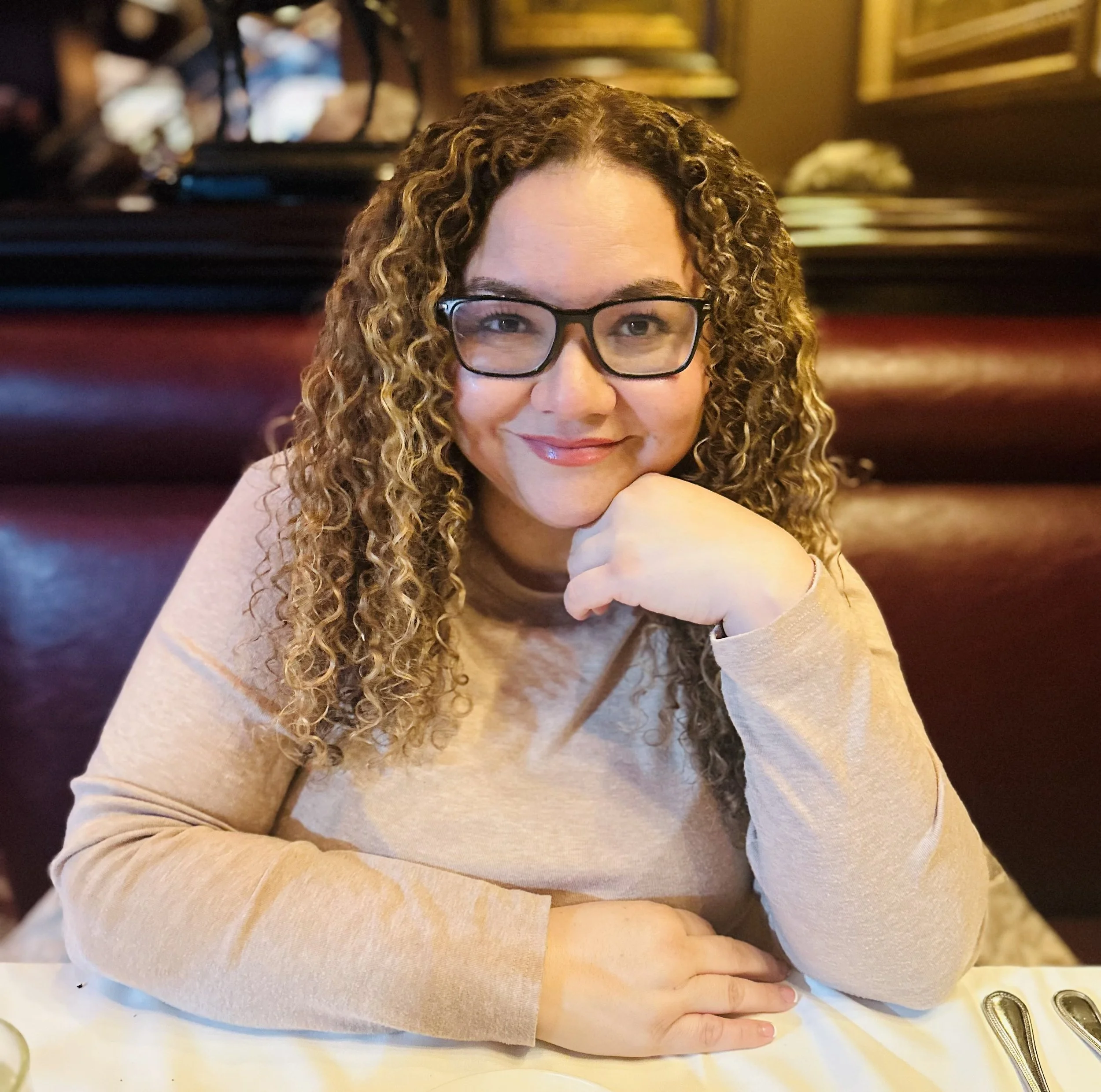 Taby with curly hair and glasses sitting at a restaurant table, smiling at the camera.