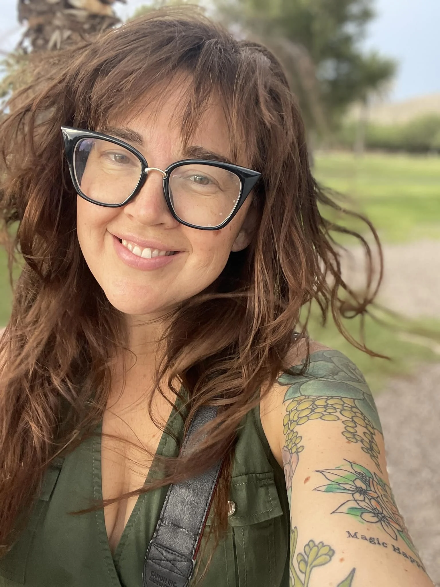Tiffany with curly brown hair wearing large black-framed glasses, a green sleeveless top, and a colorful tattoo on her left arm, smiling outdoors with a blurred natural background.
